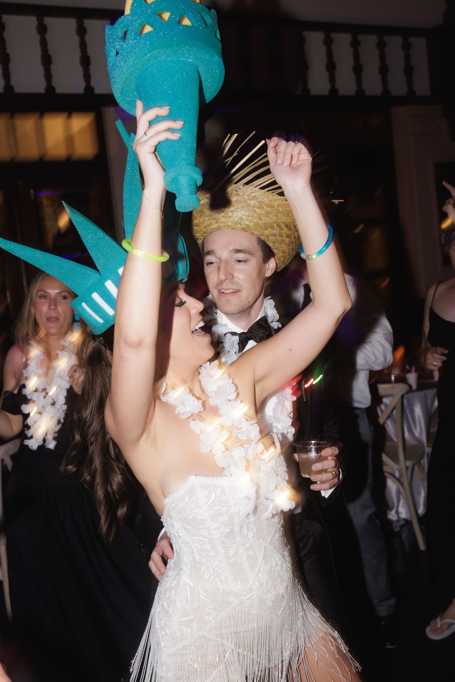Wedding party celebrates on dance floor with people waving teal foam glow sticks and dancing in formal attire.