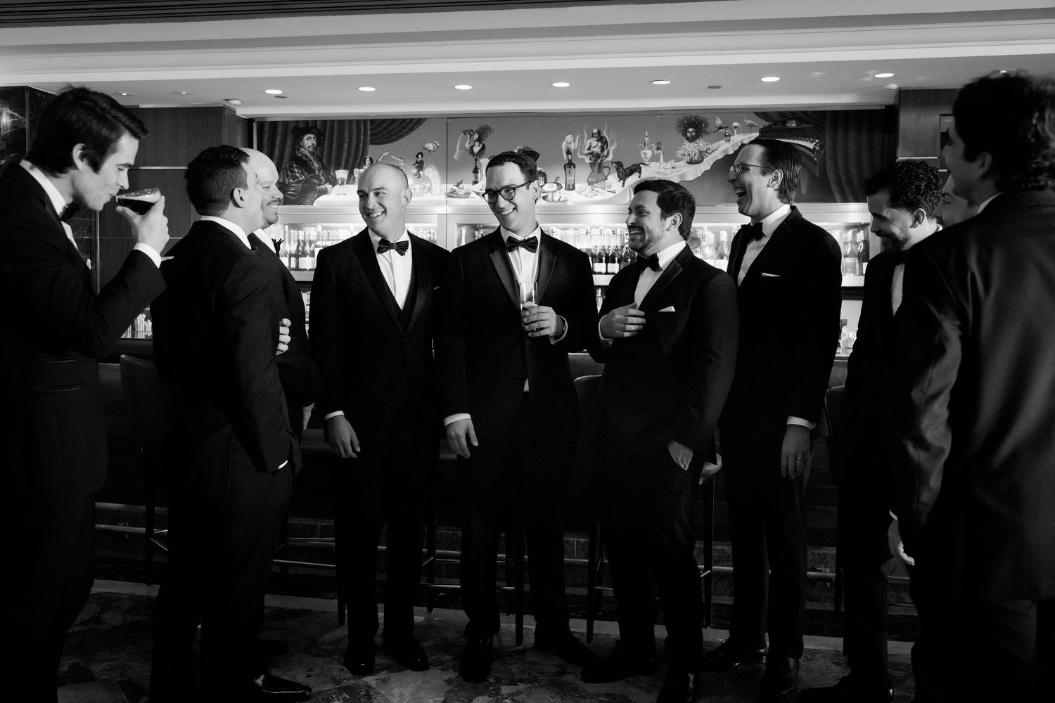 A formal wedding reception scene captured in black and white showing guests in tuxedos at a bar.