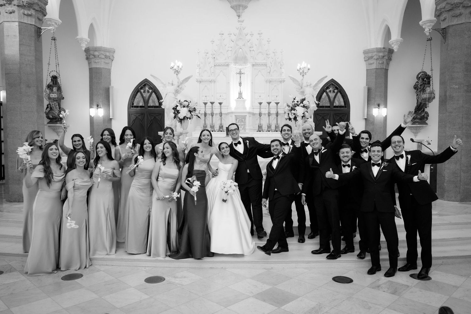 Wedding party members pose for formal group photo in an ornate church interior with high ceilings.
