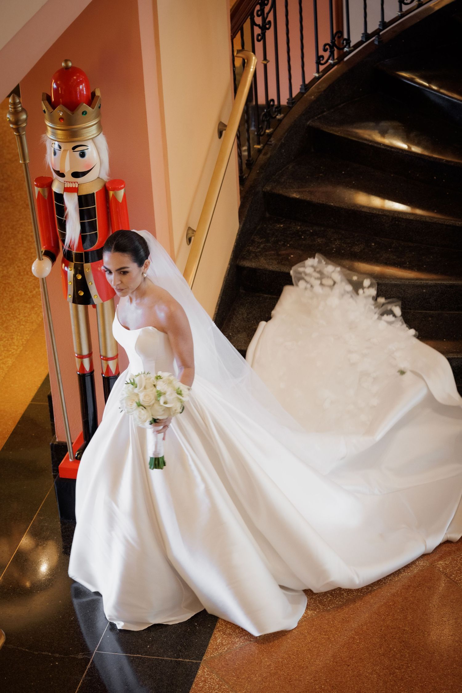 A bride in a flowing white gown stands next to a tall decorative nutcracker figure near a spiral staircase.