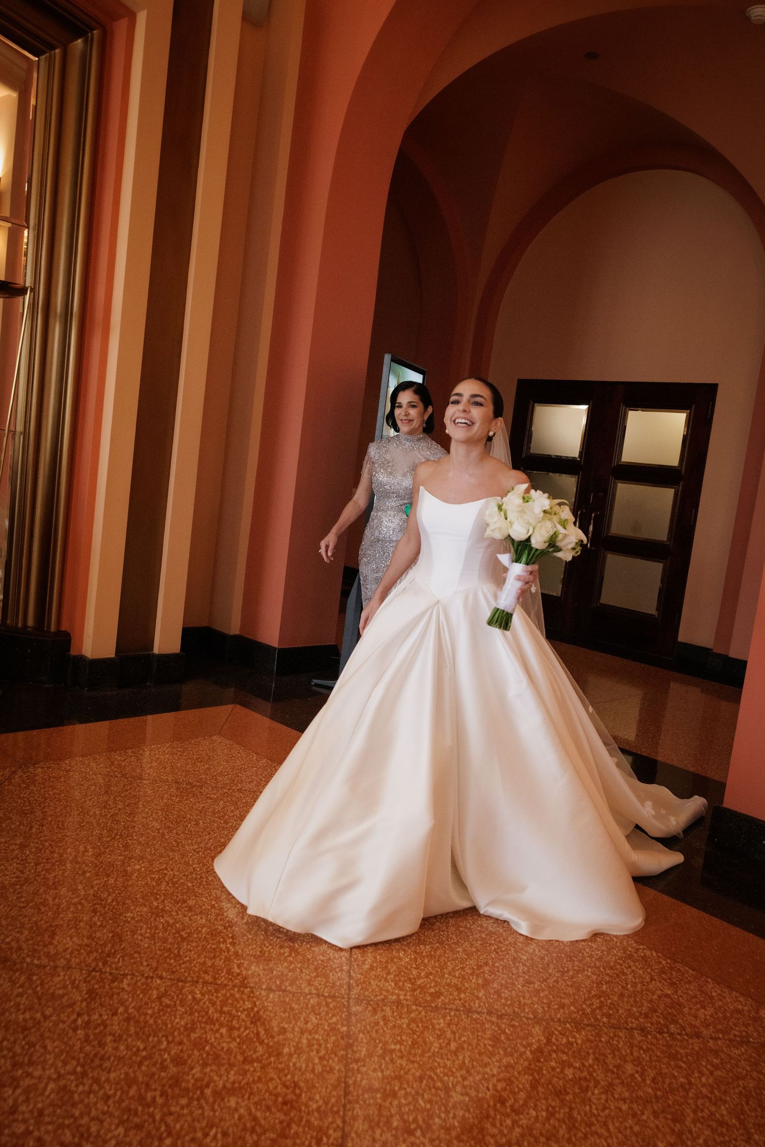 Two elegant white wedding dresses shown in a hallway with warm lighting and marble flooring.