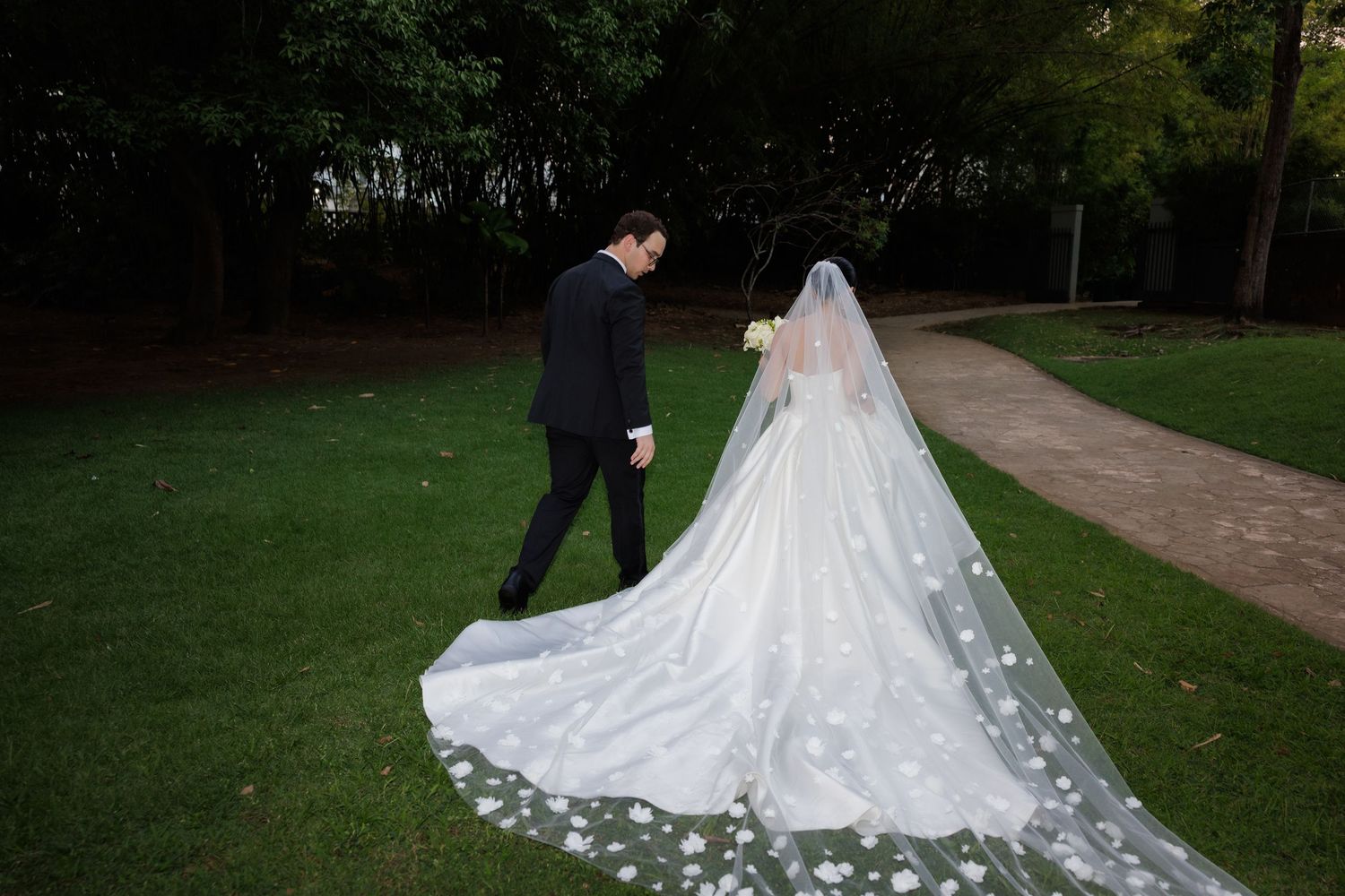 A bride in a flowing white gown with a dramatic cathedral-length train walks along a garden path.