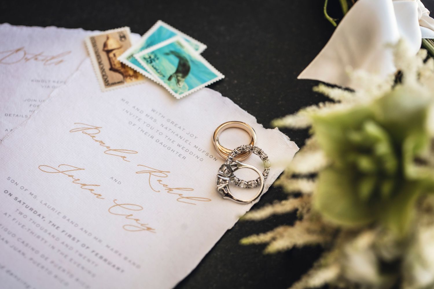 Wedding invitation with gold lettering lies beside a ring and floral arrangement on black surface.