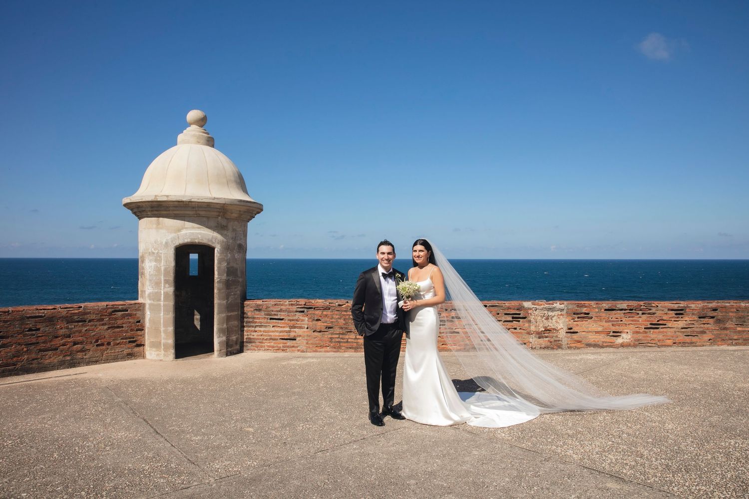 A wedding couple poses by a historic stone watchtower overlooking a scenic ocean view.