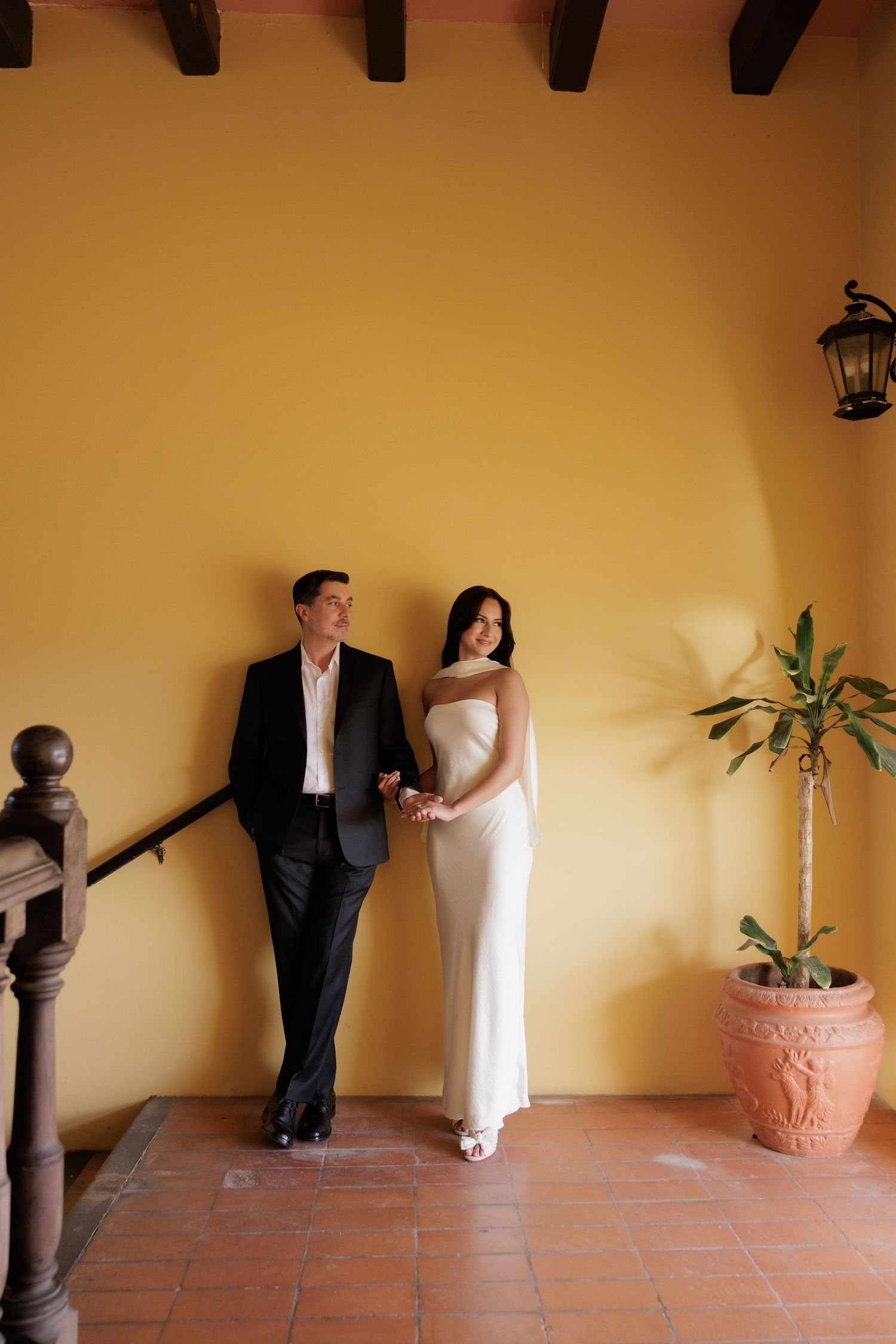 A couple poses against a warm yellow wall with terracotta tiles and Spanish colonial architecture.