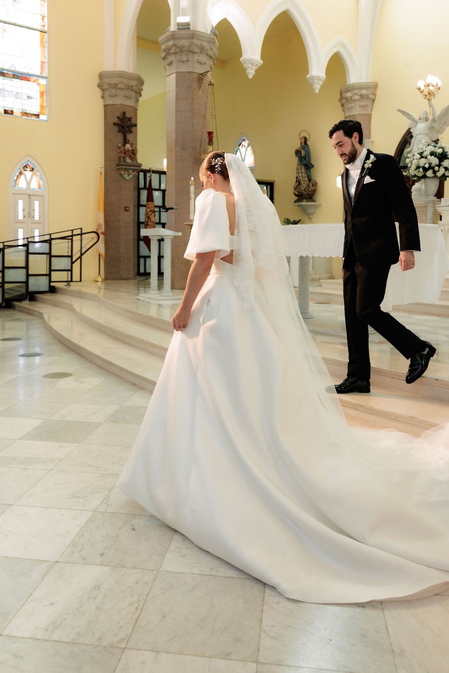 A bride in an elegant white dress with long train walks through a church with arched architecture.
