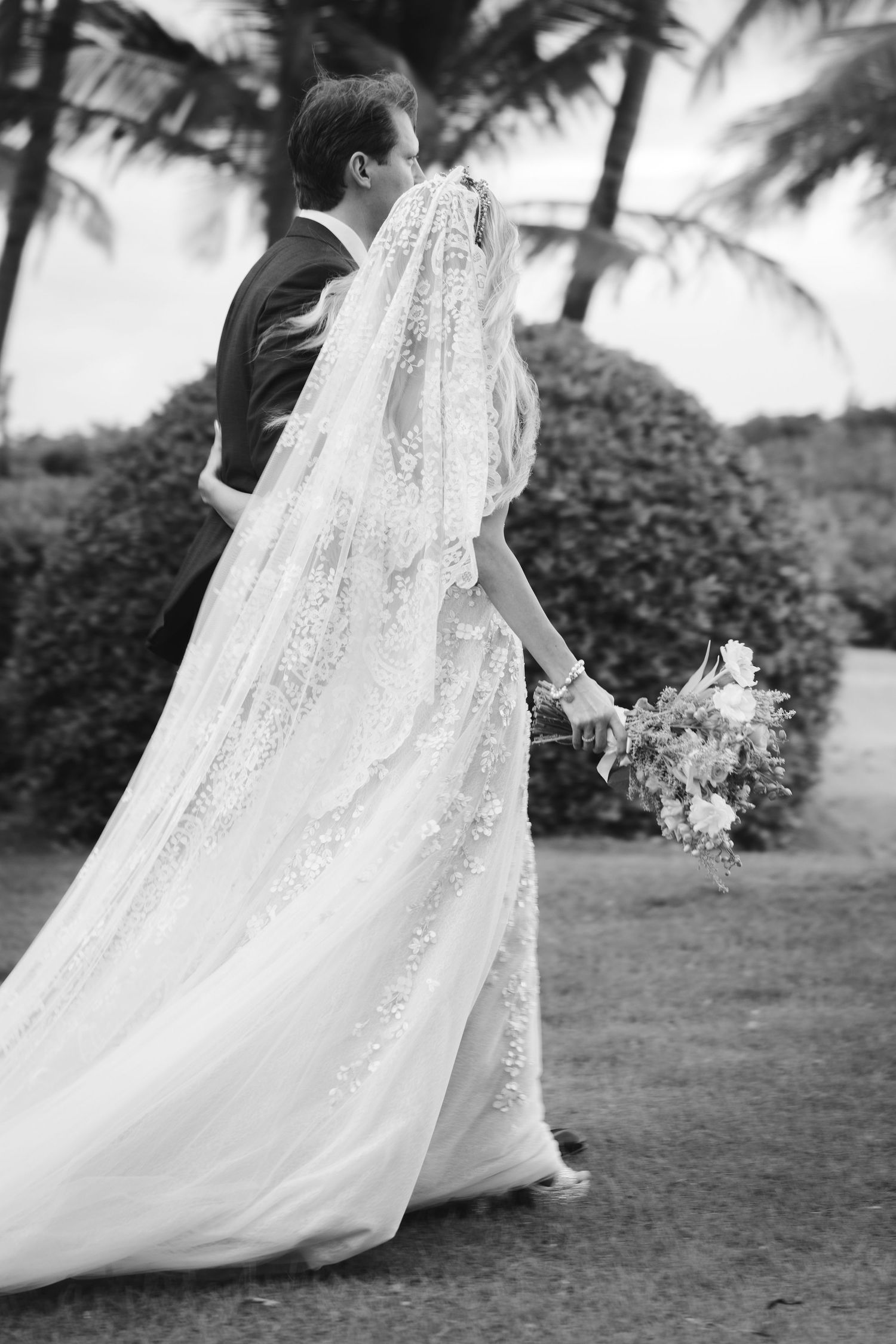 Black and white wedding photo showing couple from behind with bride in flowing lace veil and bouquet under palm trees.