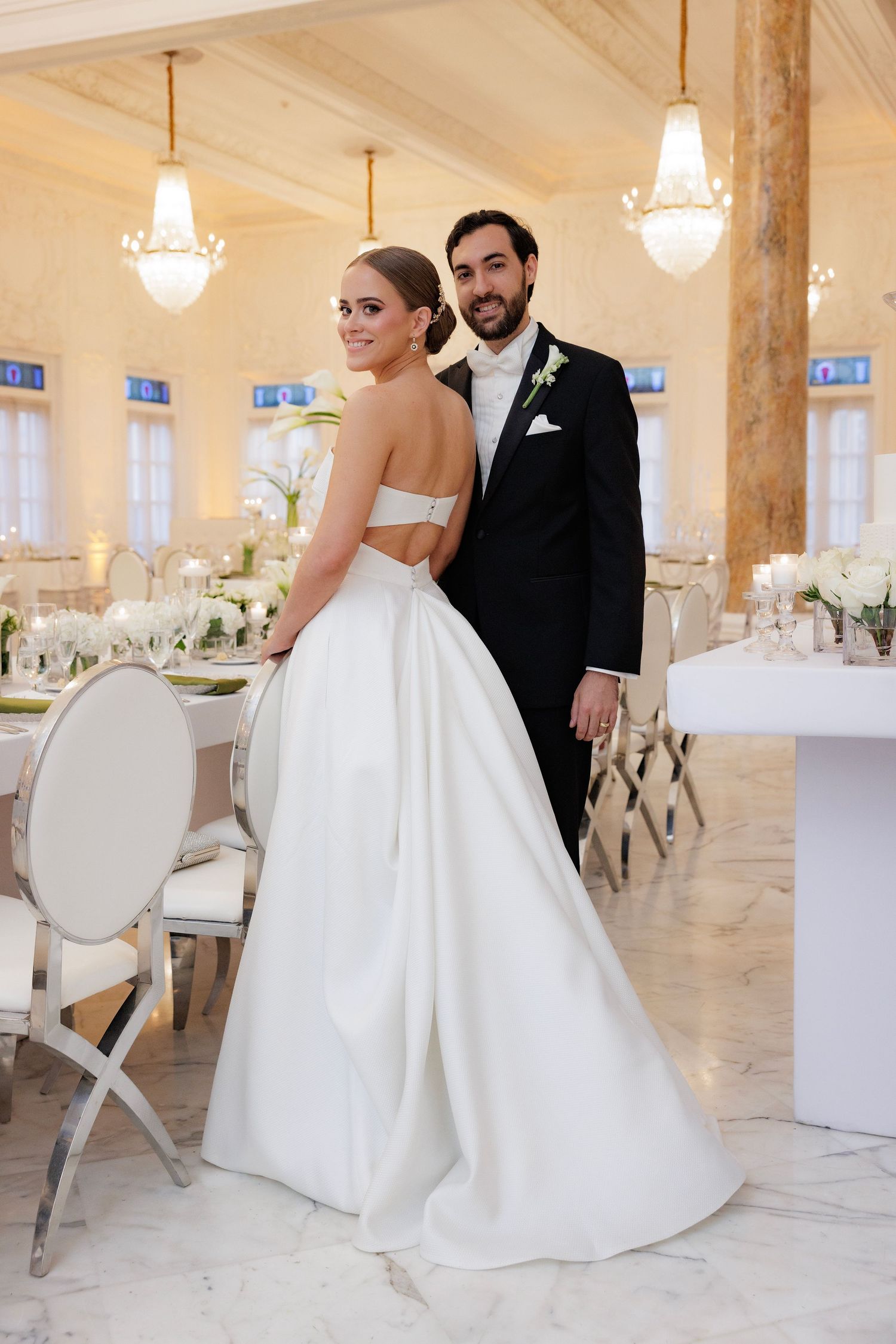 Elegantly dressed couple posing in a luxurious white reception hall with ornate chandeliers and classic decor.