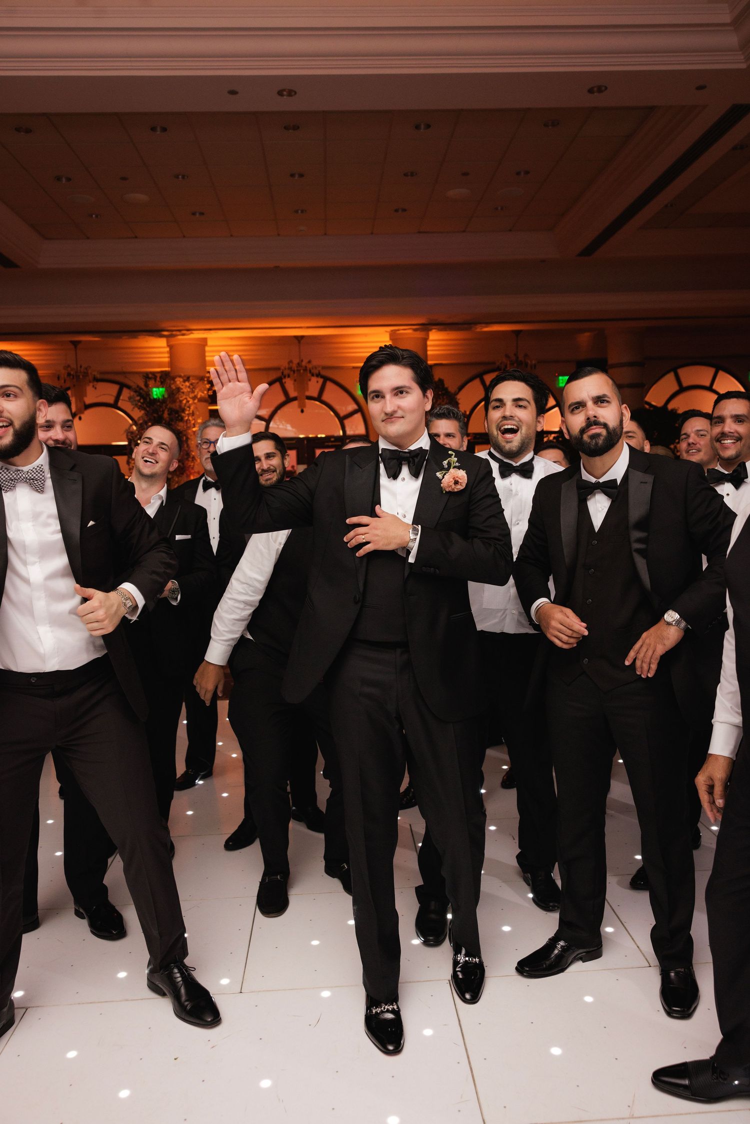 Groomsmen in black tuxedos pose together while jumping in sync on a white dance floor at a wedding reception.