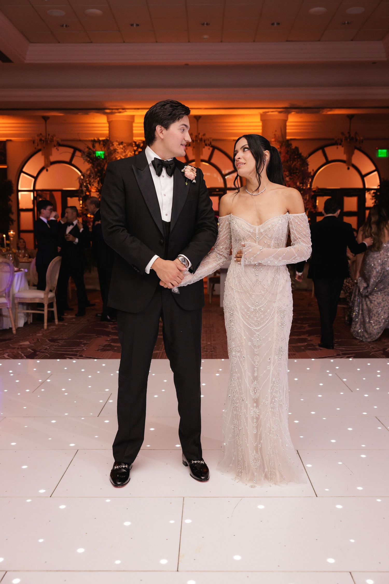 A couple shares an intimate moment on their wedding day, posed on a white dance floor with ornate arches in the background.