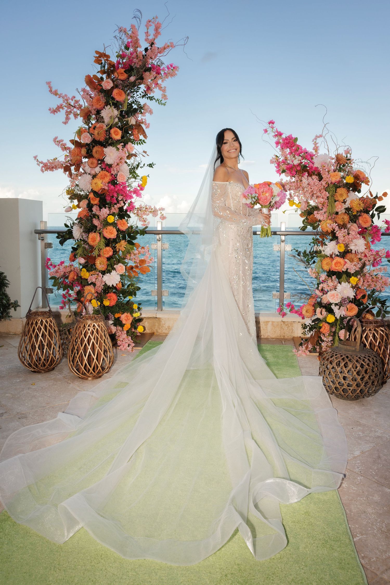 Flowing wedding gown train spread across seaside venue with vibrant coral and pink floral arrangements.
