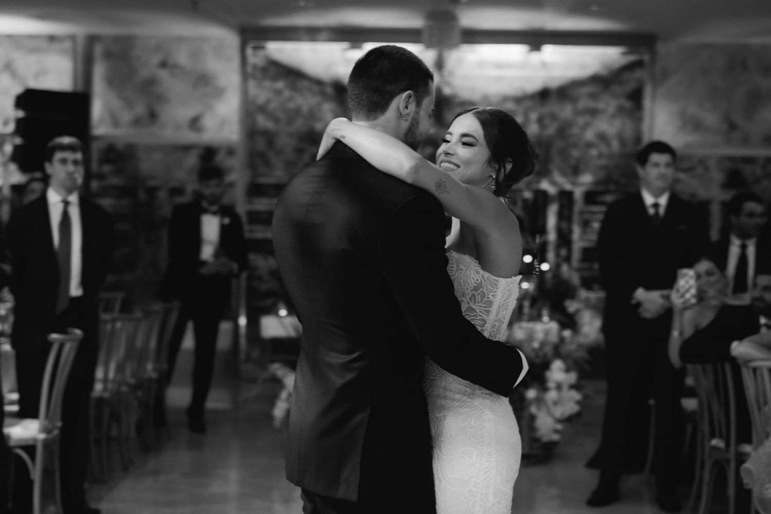 A tender black and white photo of newlyweds sharing their first dance, surrounded by guests at their reception.