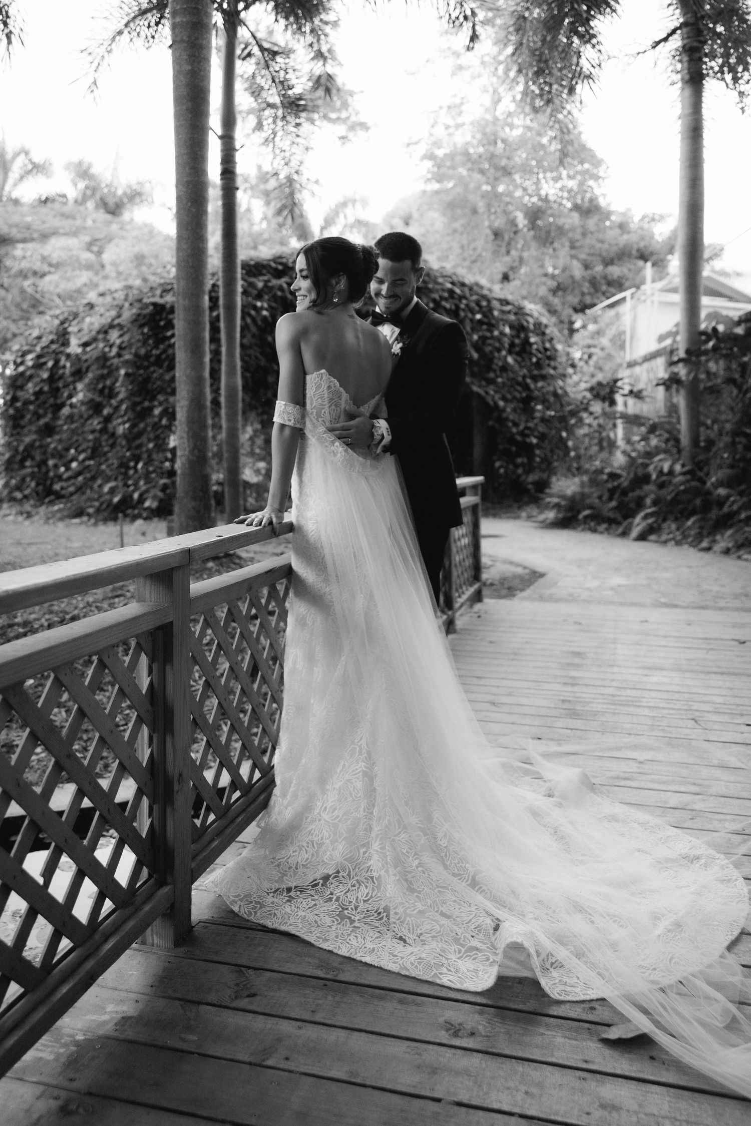 Black and white photo of couple on wooden deck with bride's lace train in focus.