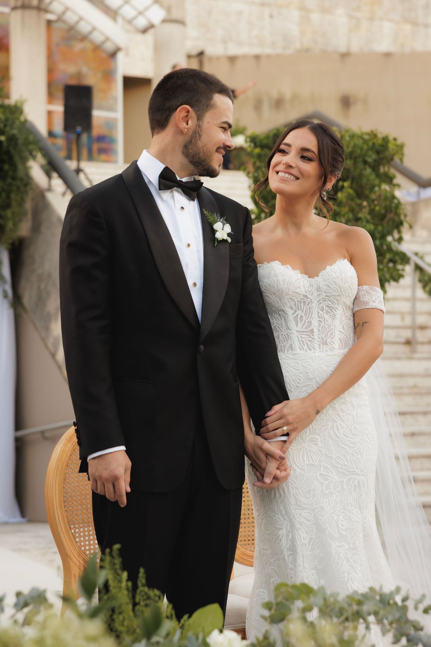 Elegant wedding couple standing beside vintage rattan chairs surrounded by white floral arrangements outdoors.