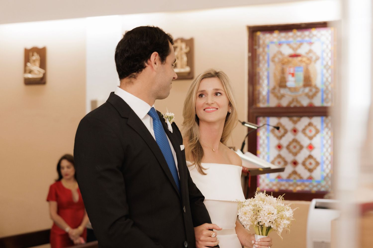 A vintage-style wedding couple leans in for a kiss in a warm-toned hallway with framed artwork on the wall.