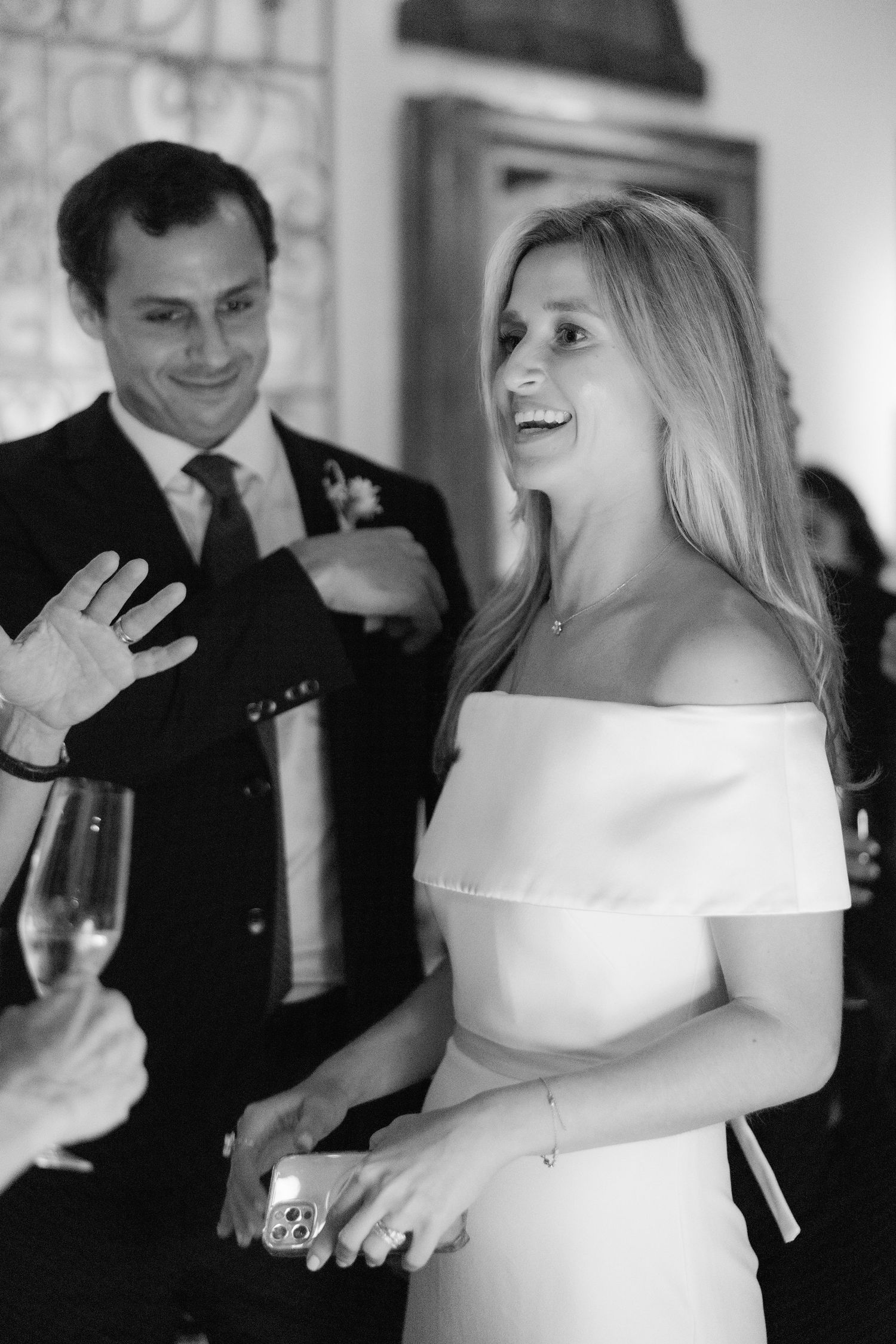 A candid black and white wedding photo showing a bride in an off-shoulder dress laughing during a reception celebration.