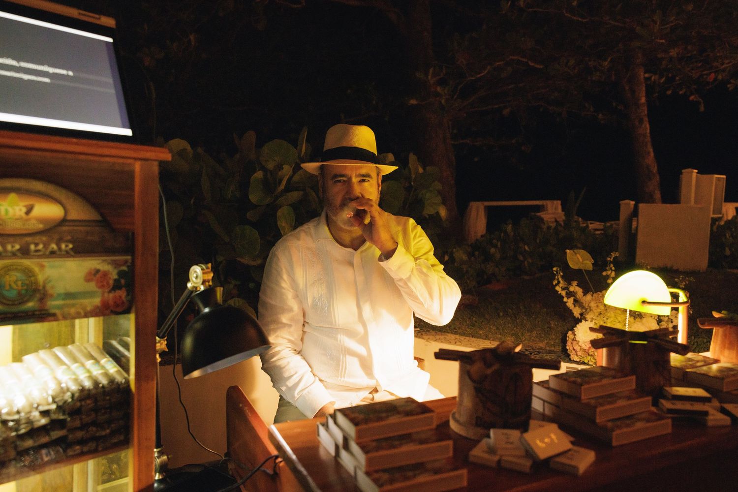Vendor in a yellow-lit stall at a nighttime market wearing a white shirt and hat while tending to wooden boxes.