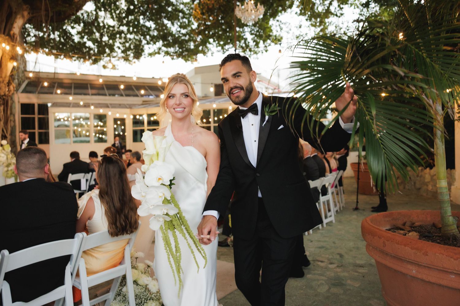 A couple walks down the aisle holding hands at their outdoor wedding with palm trees and chairs in view.