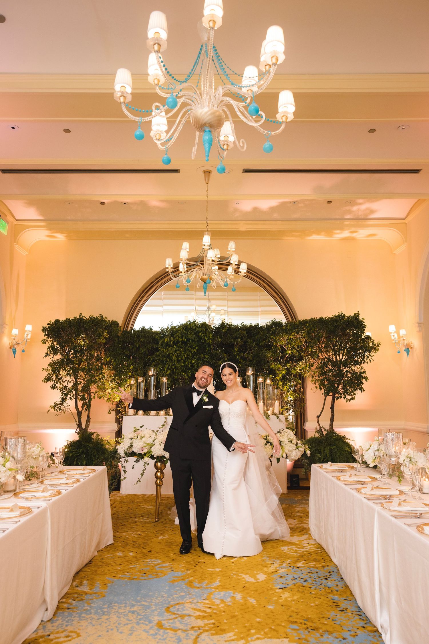 Elegant wedding reception venue features crystal chandeliers, greenery arch, and gold carpeting between white-clothed tables.