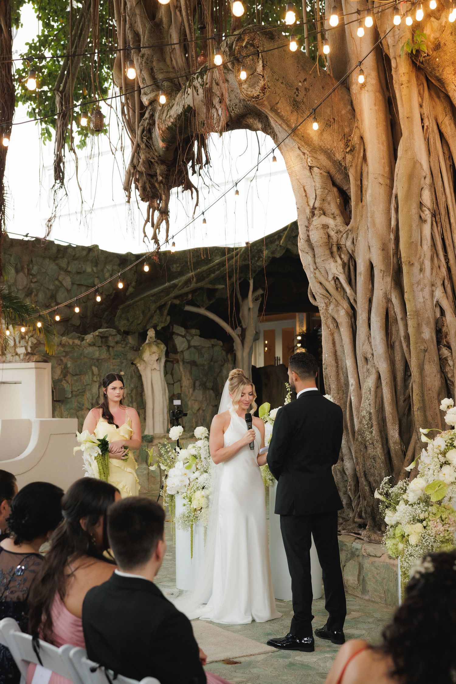 A romantic outdoor wedding ceremony beneath illuminated banyan trees with string lights and white floral decorations.