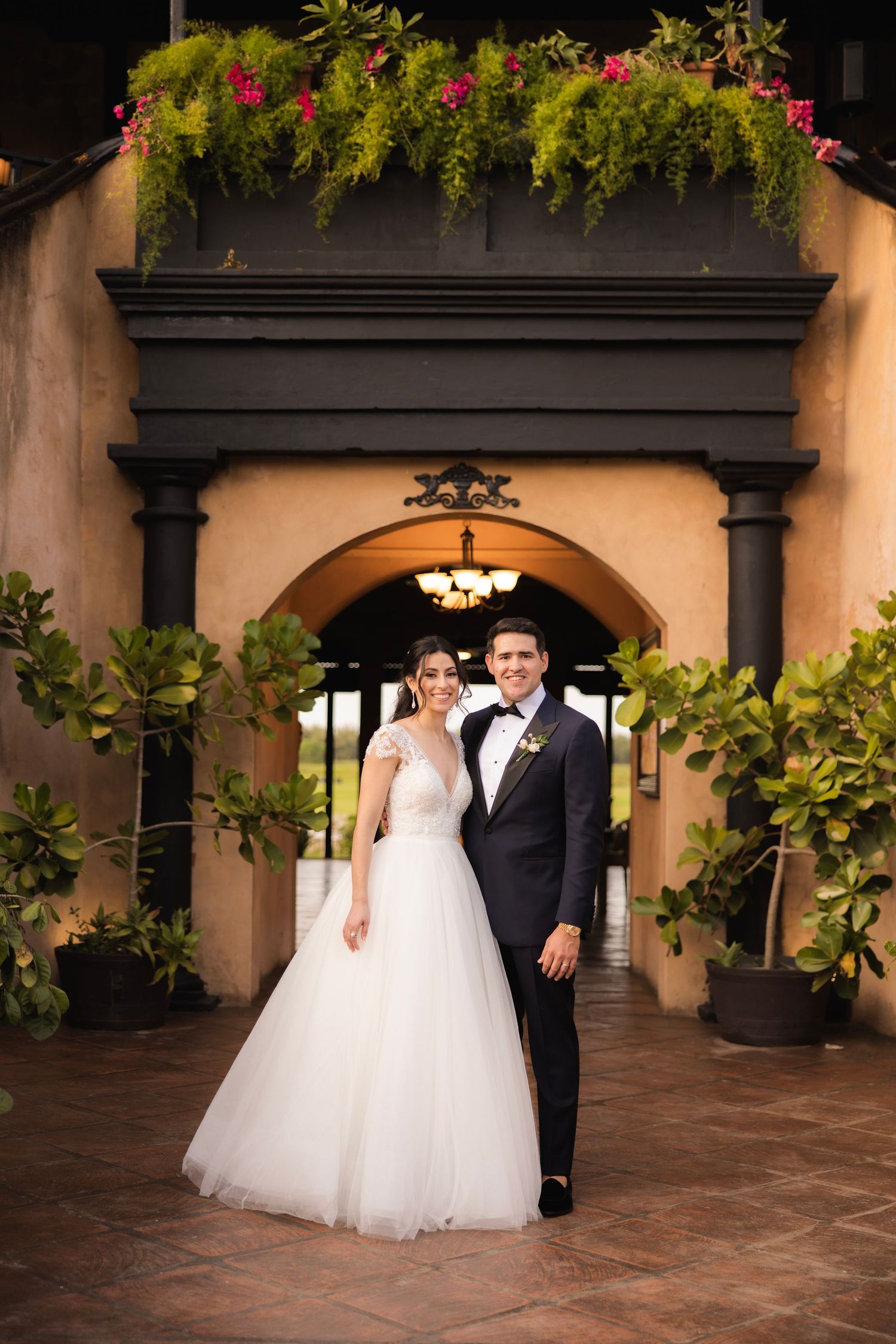 Wedding couple poses under an elegant Spanish-style archway decorated with hanging pink flowers and greenery.