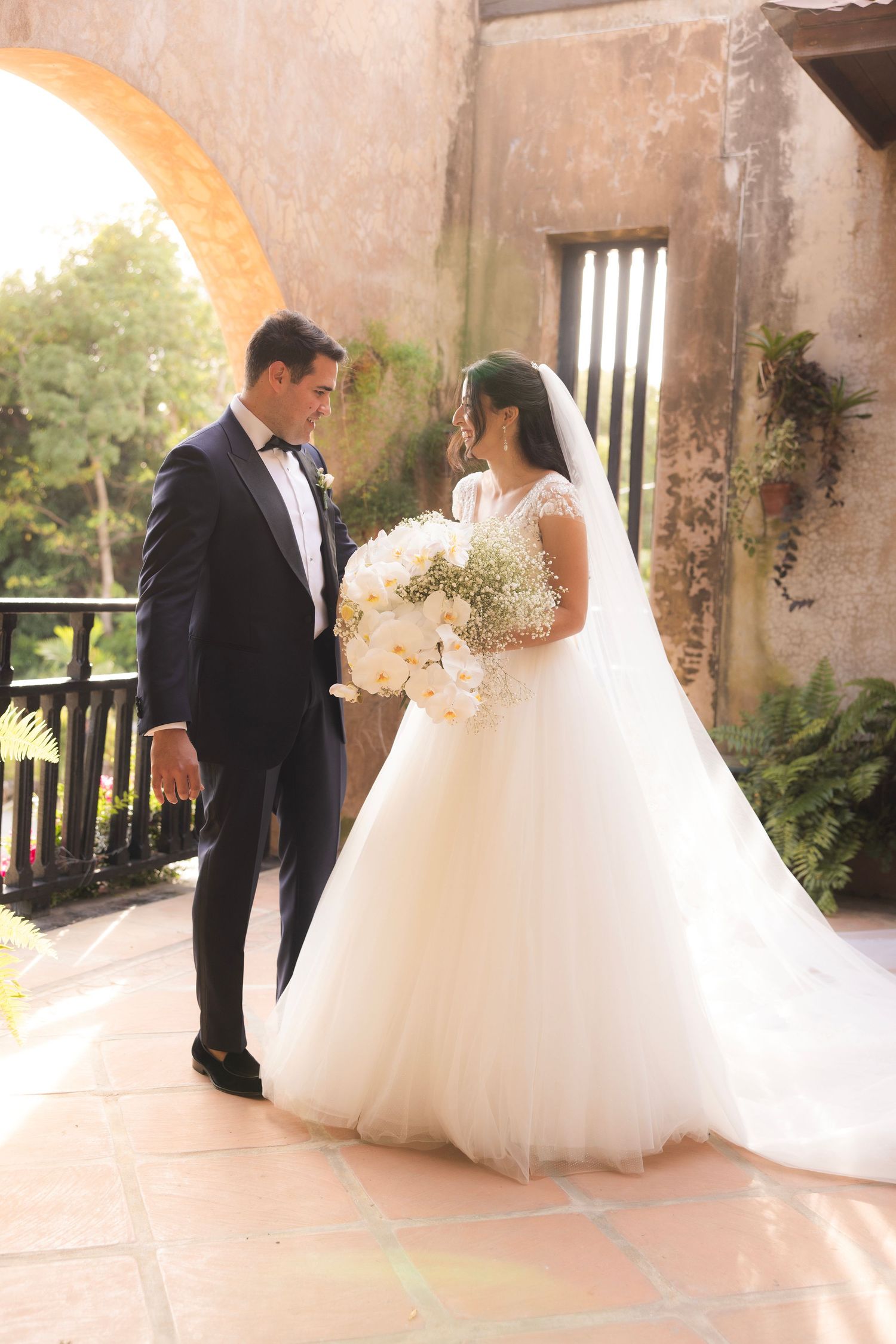 Romantic wedding couple posing on rustic balcony in sunlit courtyard.