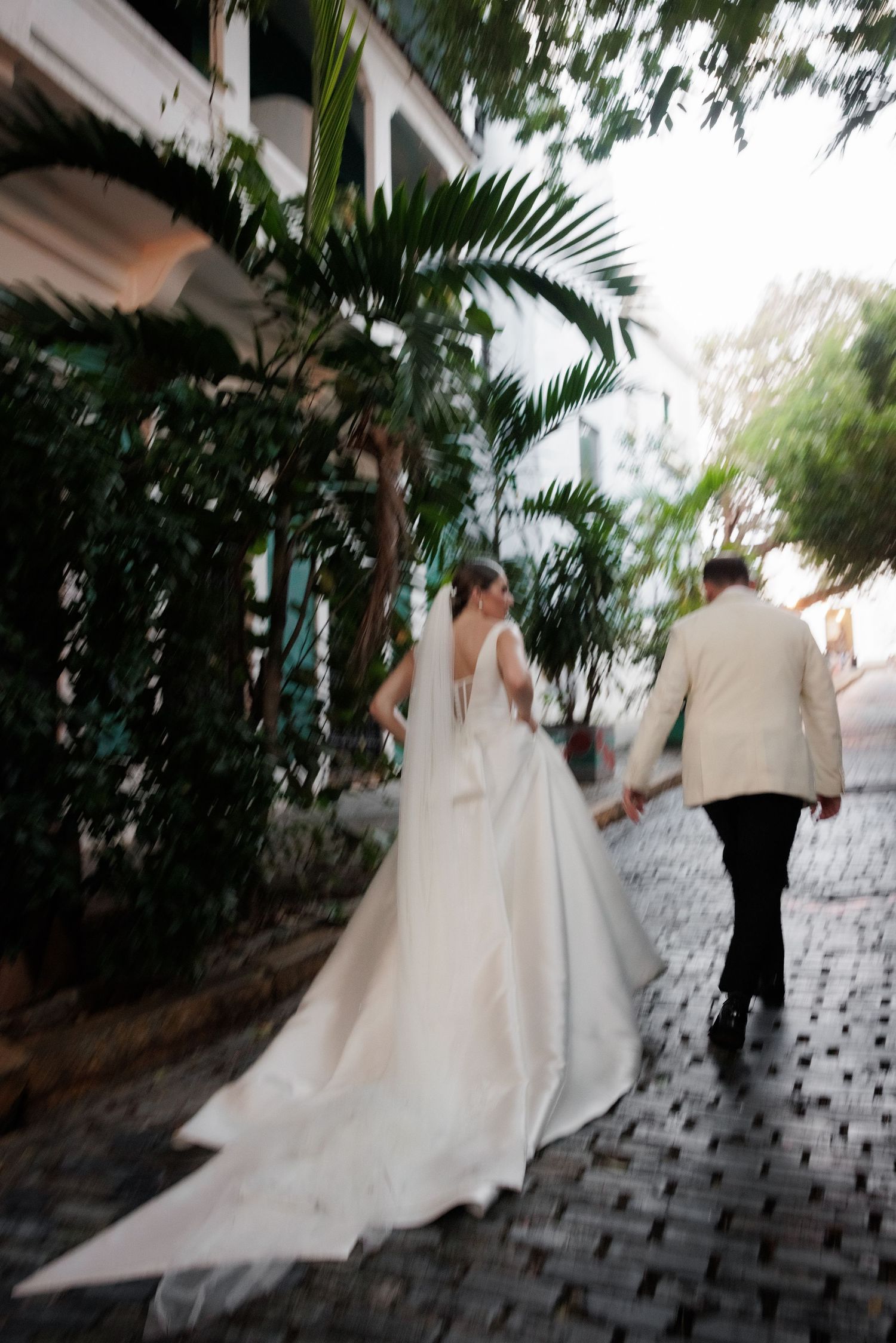 Wedding couple walking along a palm-lined path with flowing white dress trailing behind.