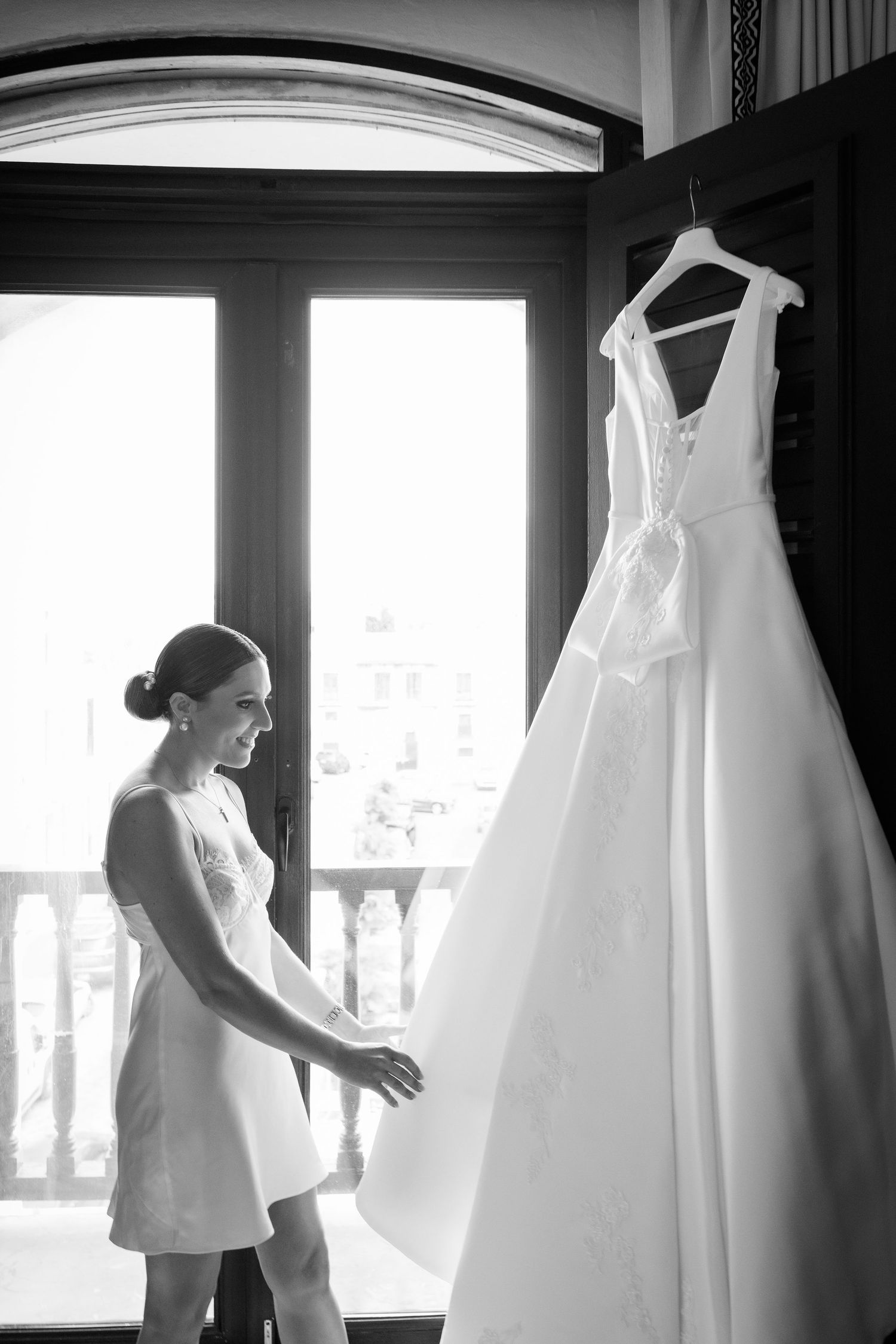 A white wedding dress hangs in a sunlit room while someone in a short white dress admires it.