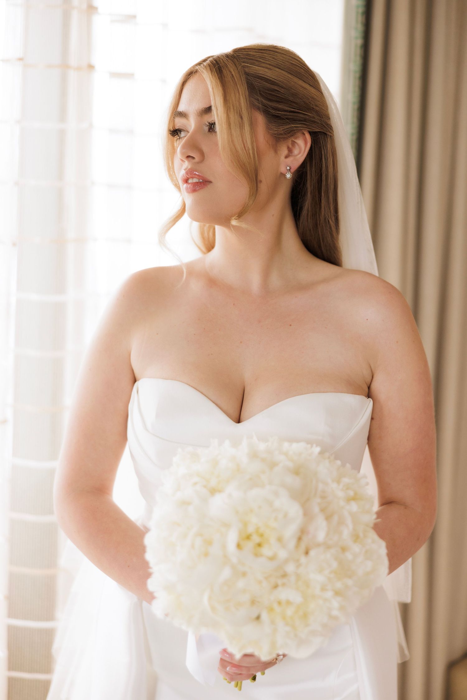 Elegant bride in strapless white gown holding a bouquet of white peonies against a window.