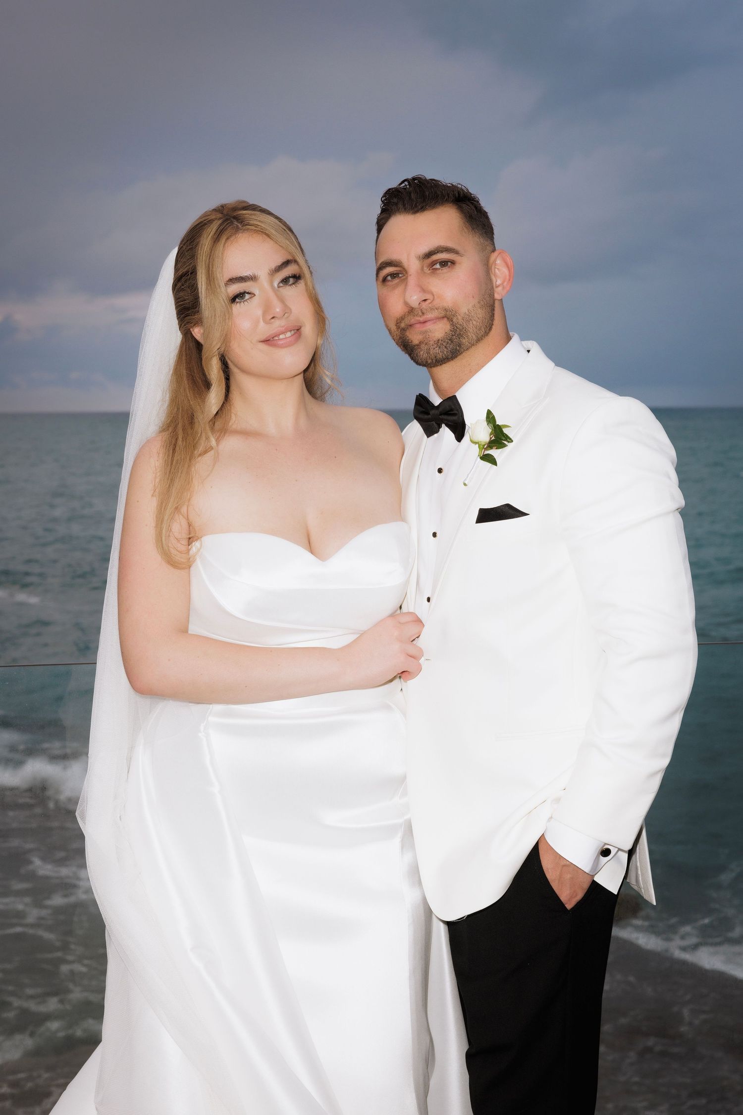 A couple poses for their wedding portrait on a beach at dusk, wearing white formal attire against an ocean backdrop.