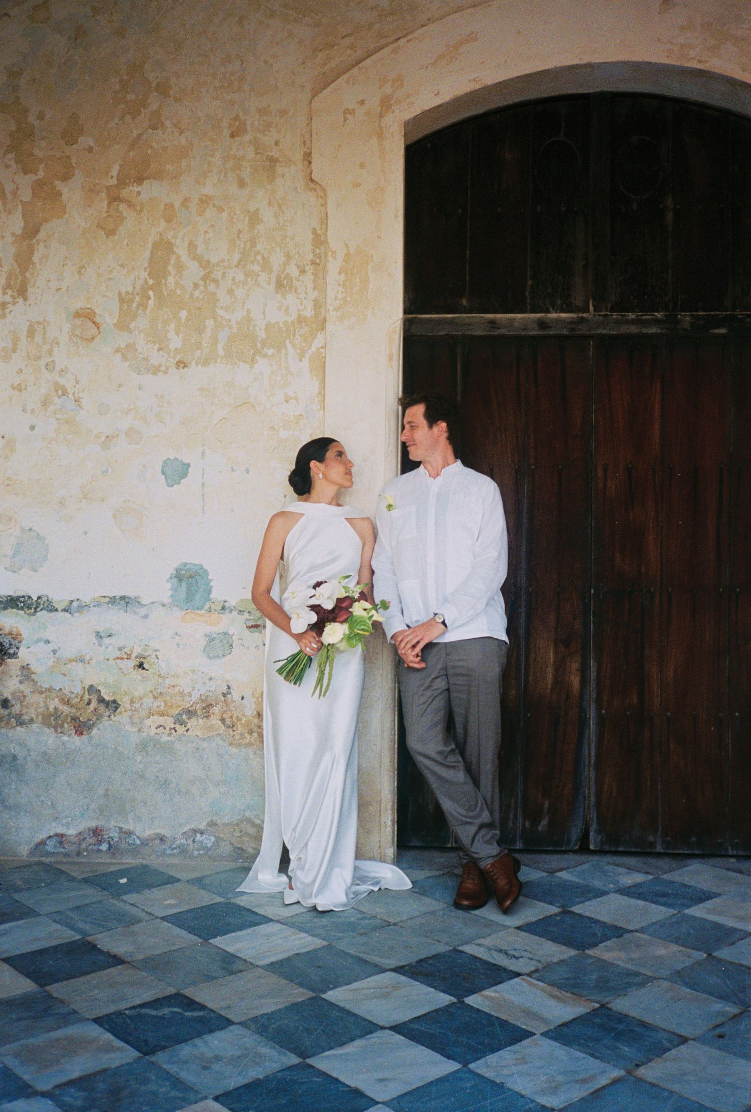 Bride and groom share a romantic moment by a rustic wooden door, she holds a bouquet in her elegant white gown.