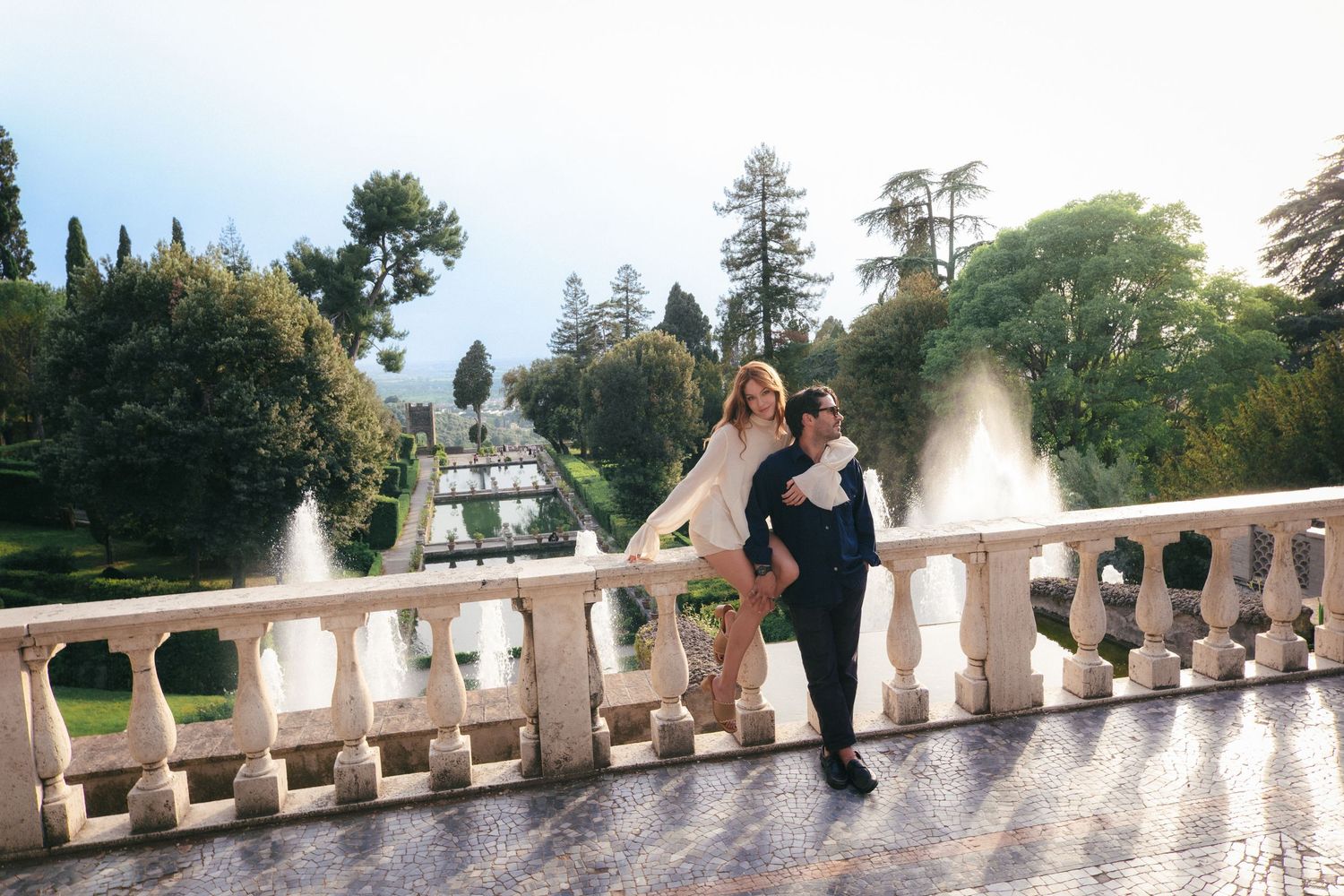 Couple posing on elegant stone balcony with fountain garden backdrop at a scenic European villa.