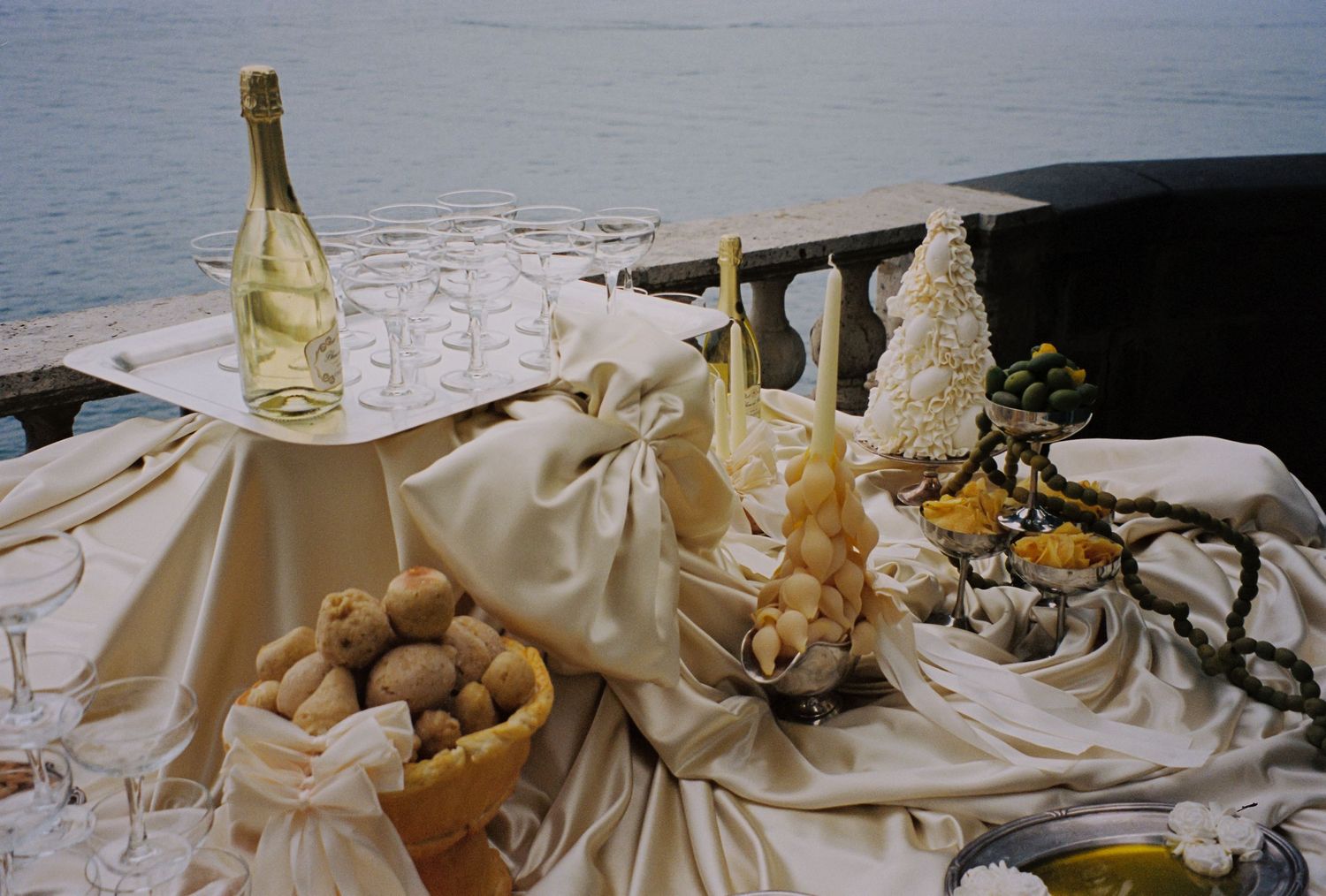 Elegant outdoor table with champagne bottle, glasses, bread, and fruits draped in white cloth by the sea.