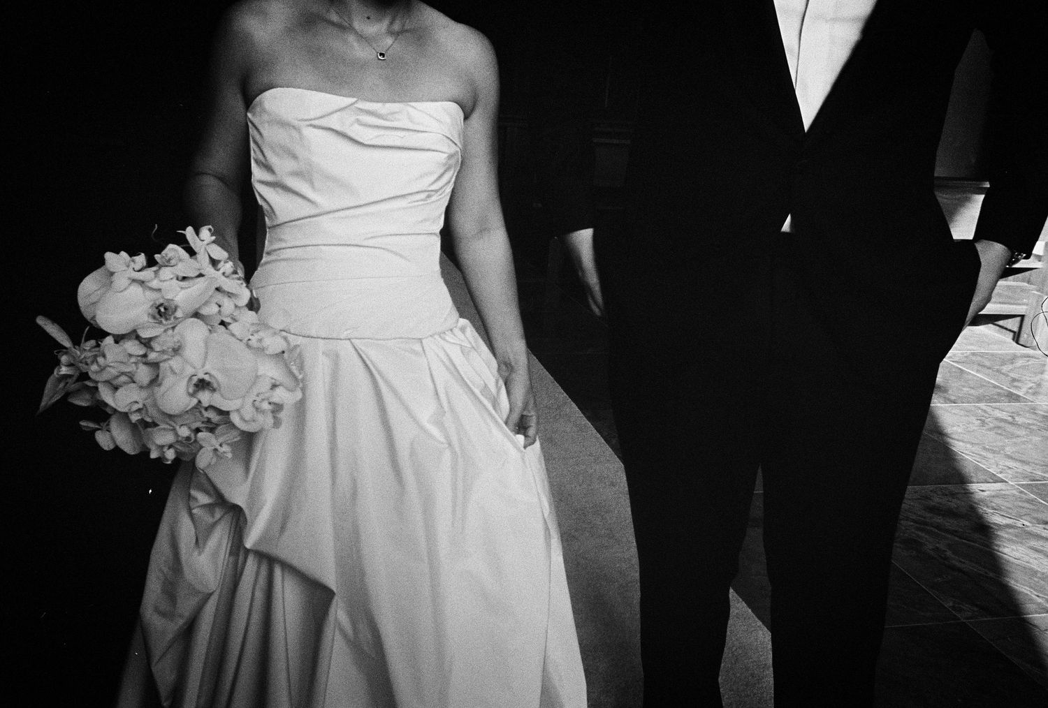 Black and white photo of bride in strapless gown holding bouquet, standing beside groom in dark suit.