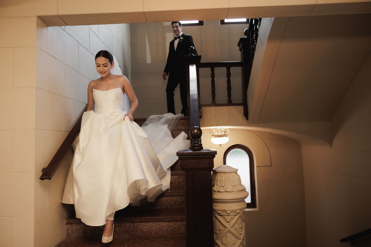 A bride in a white wedding gown walks down a beige hallway with a dramatic flowing train behind her.