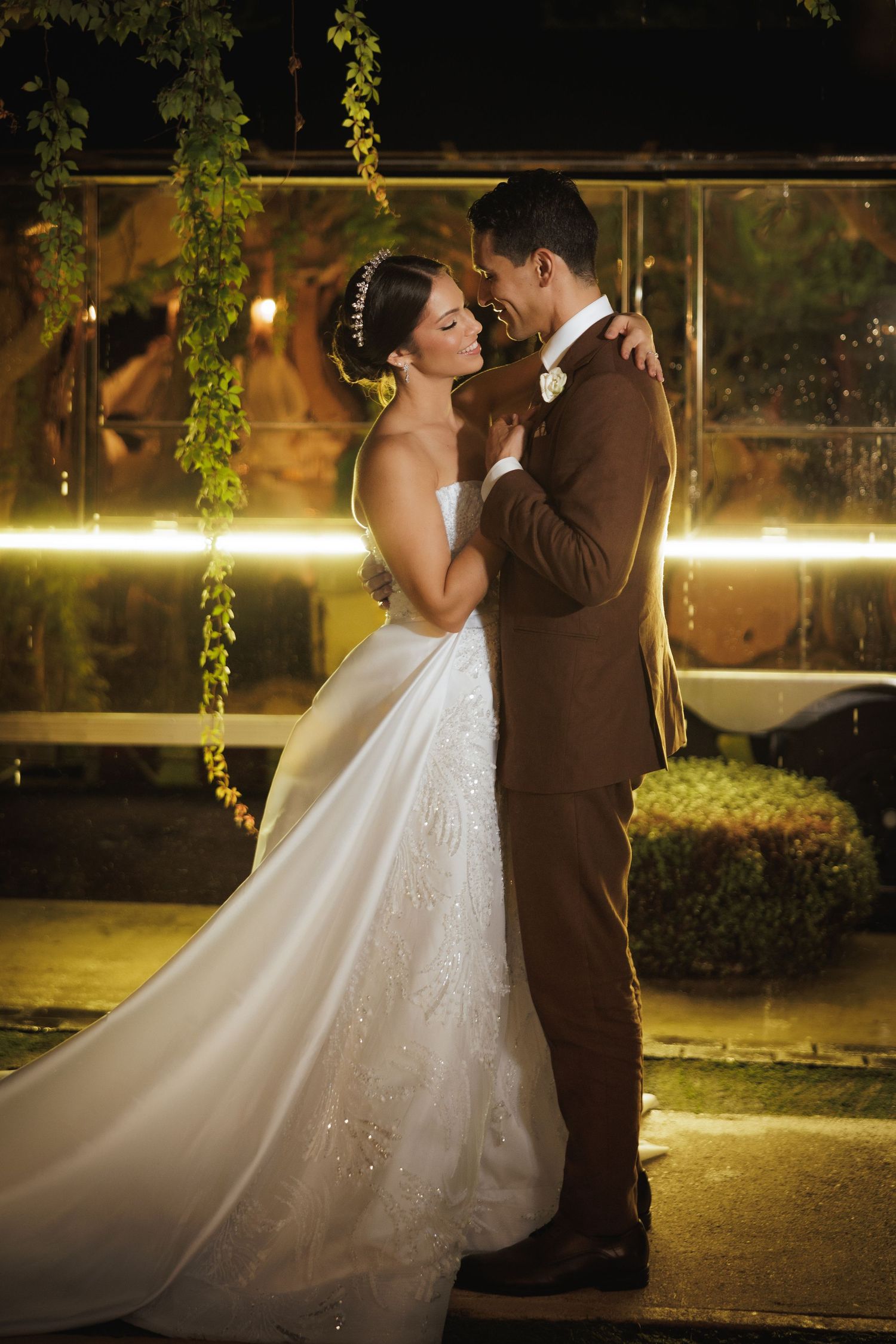 Romantic wedding couple shares an intimate moment under glowing lights and hanging greenery at night.