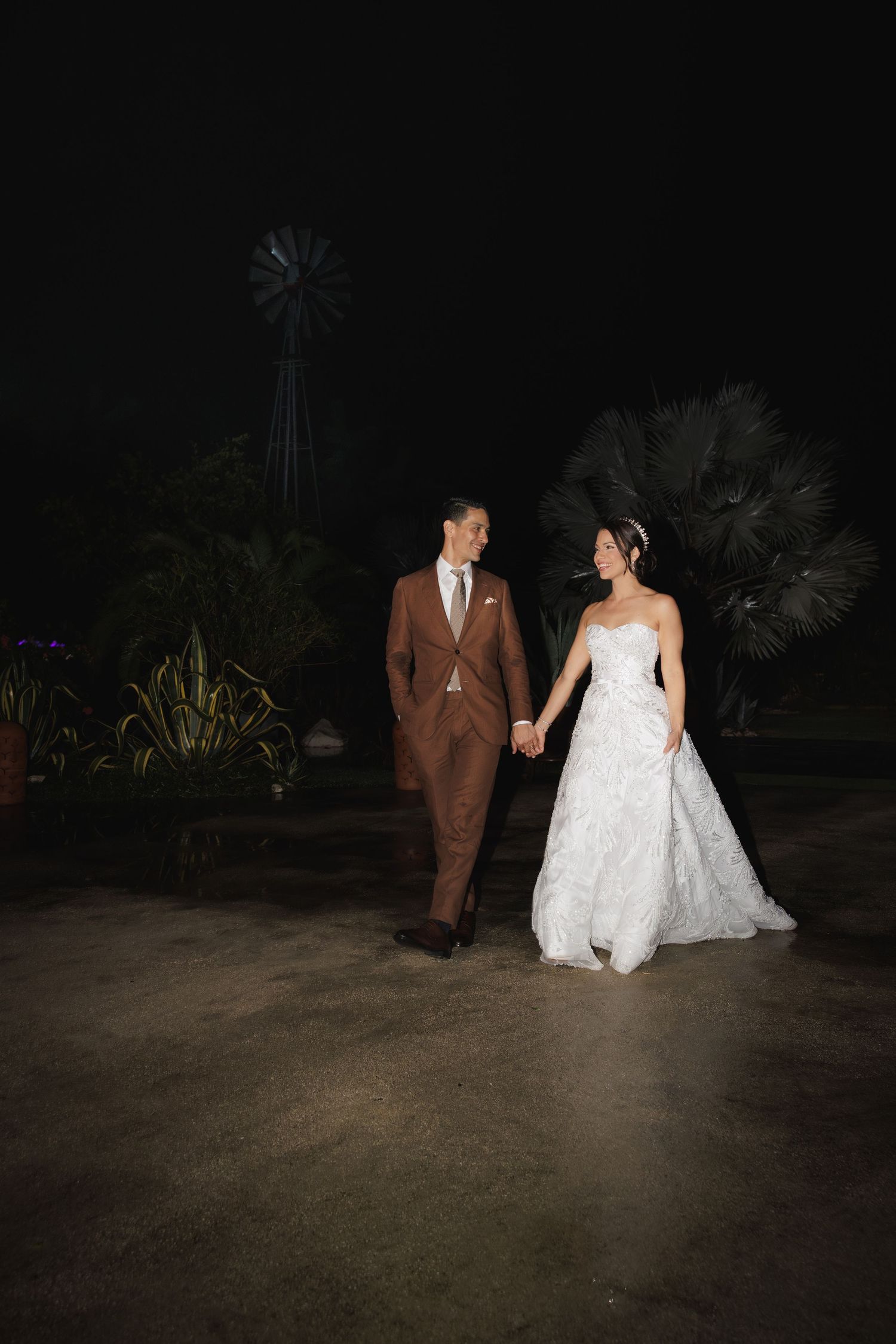 A couple in wedding attire walking together at night on a garden path.