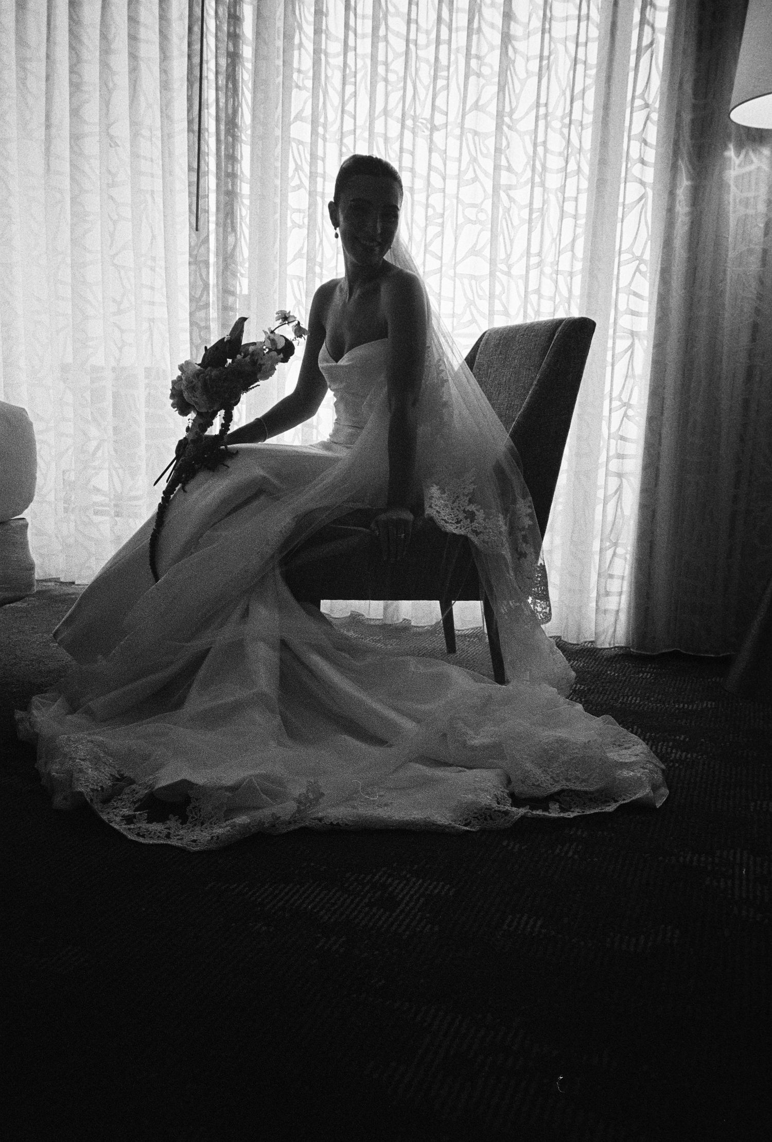 Black and white photo of a bride in a full gown and veil seated in a chair holding a bouquet.