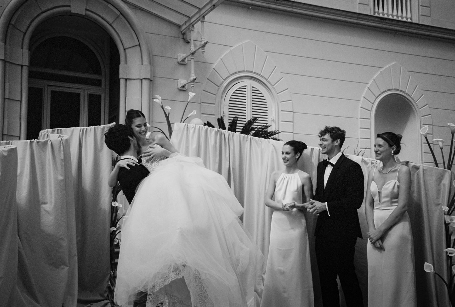 Black and white wedding photo with bride being lifted near hanging white fabric outdoors.