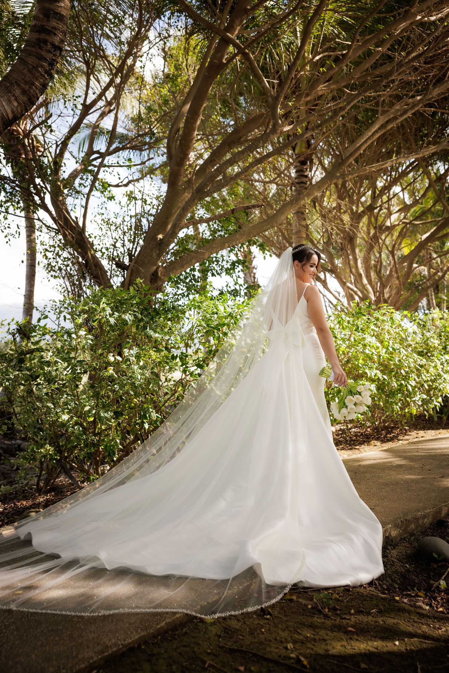 A flowing white wedding gown with long train and veil photographed in a scenic garden path with sunlit trees.