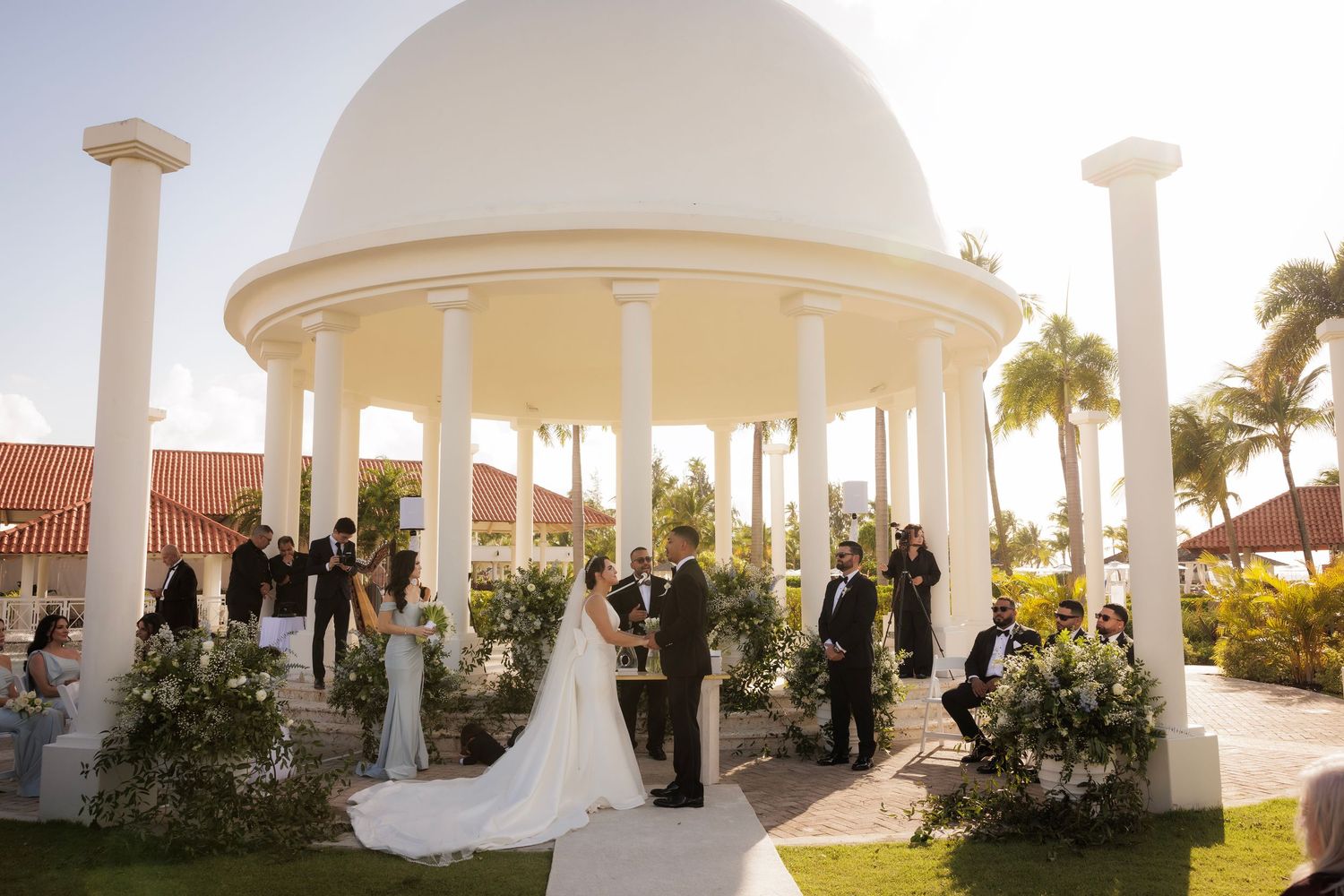 Wedding ceremony setup at a beachside gazebo with white columns and a panoramic ocean view at sunset.