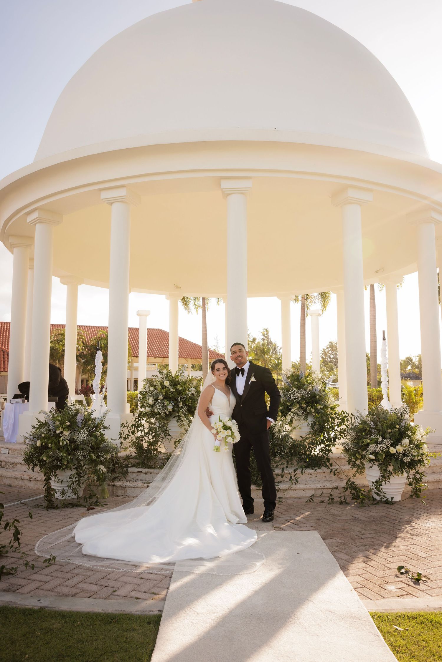 A couple poses under an elegant white columned pavilion on a sun-drenched wedding day.