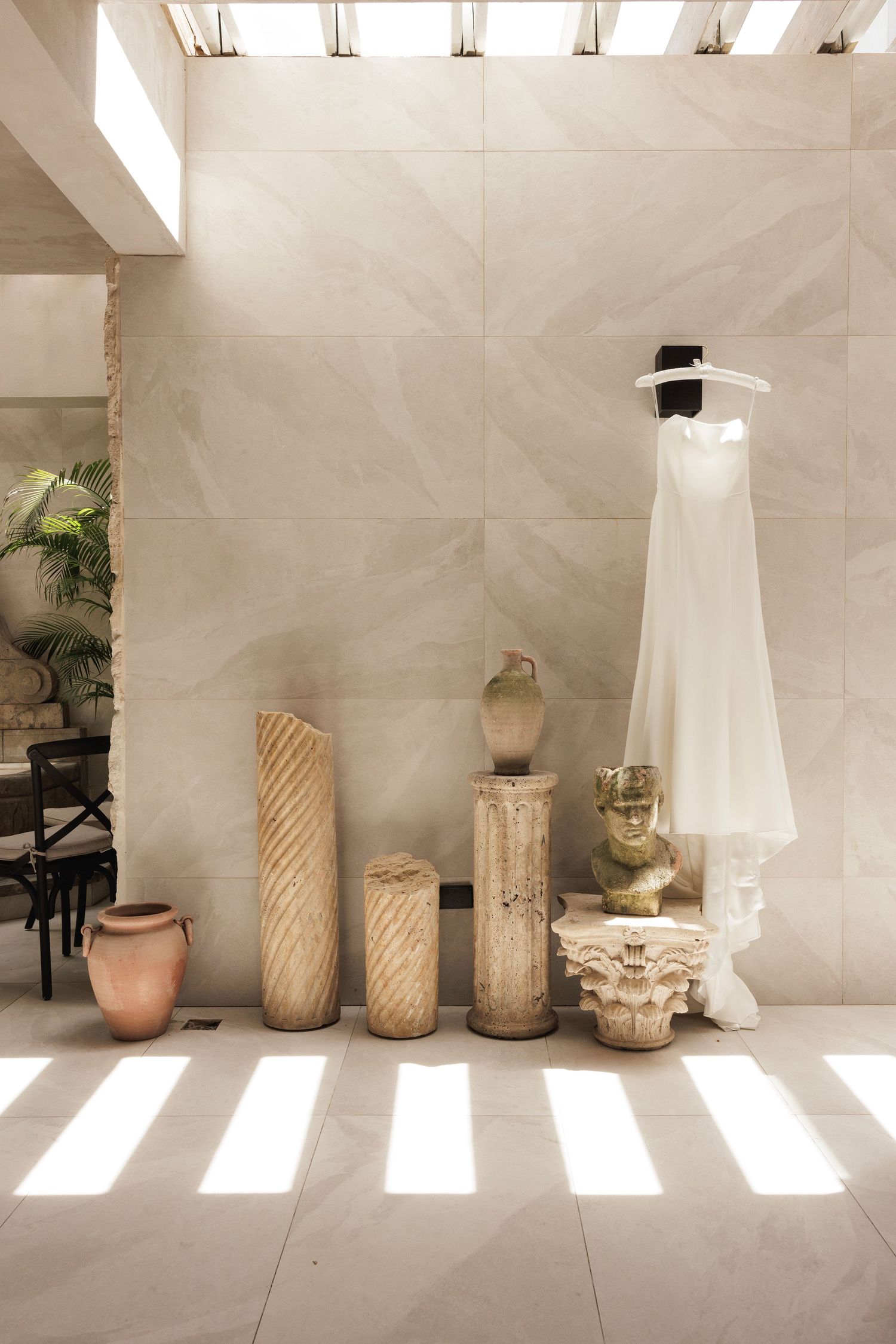 A white wedding dress hangs in an elegant space decorated with rustic wooden columns and pottery against beige walls.