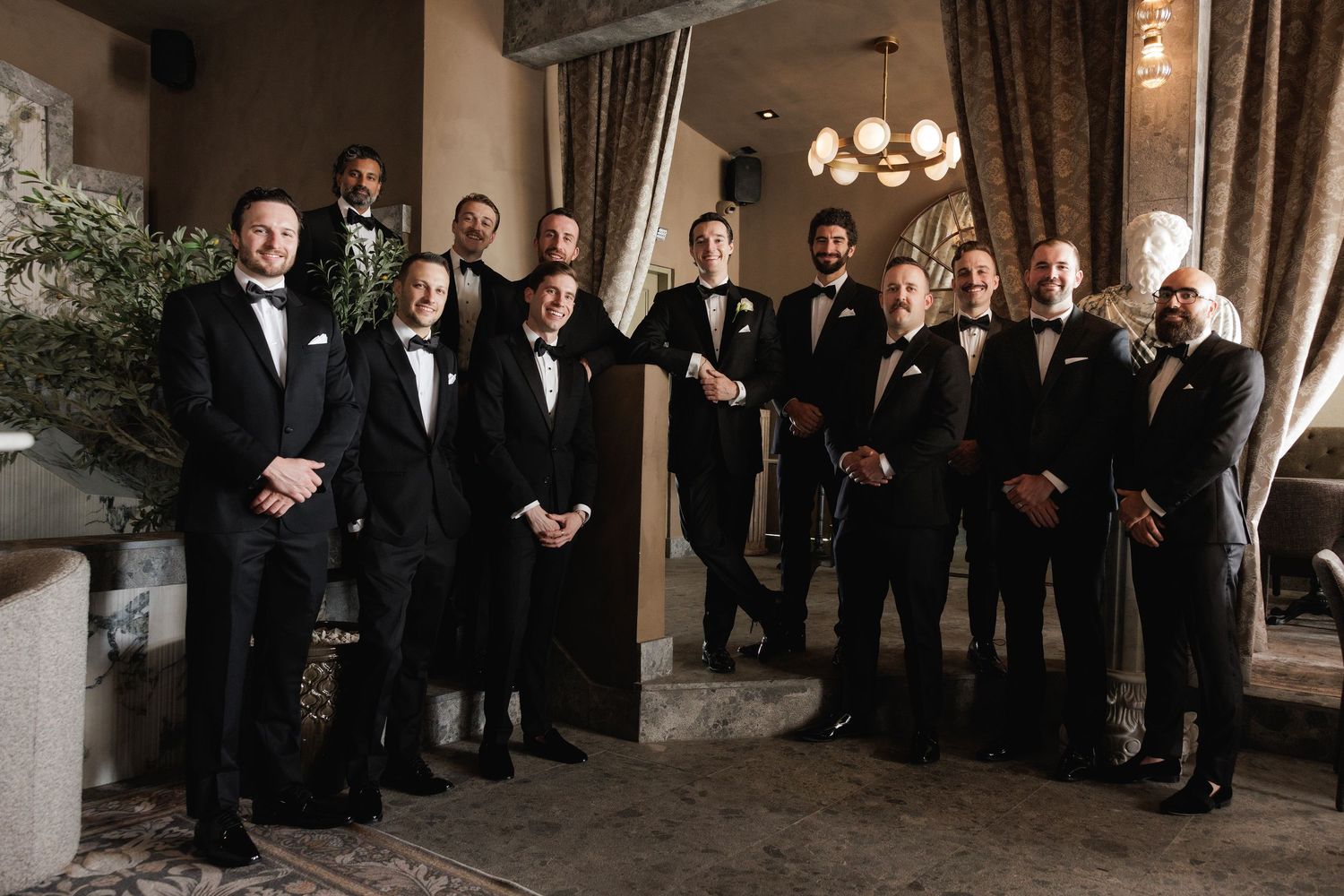 A large formal wedding party in black and white attire poses in an elegant indoor venue during a celebration.