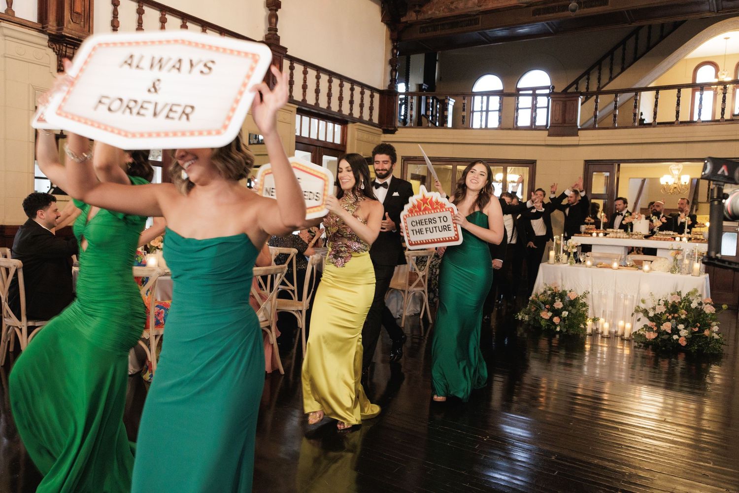 Wedding guests in green and yellow dresses hold up fun celebration signs during a reception in an elegant banquet hall.