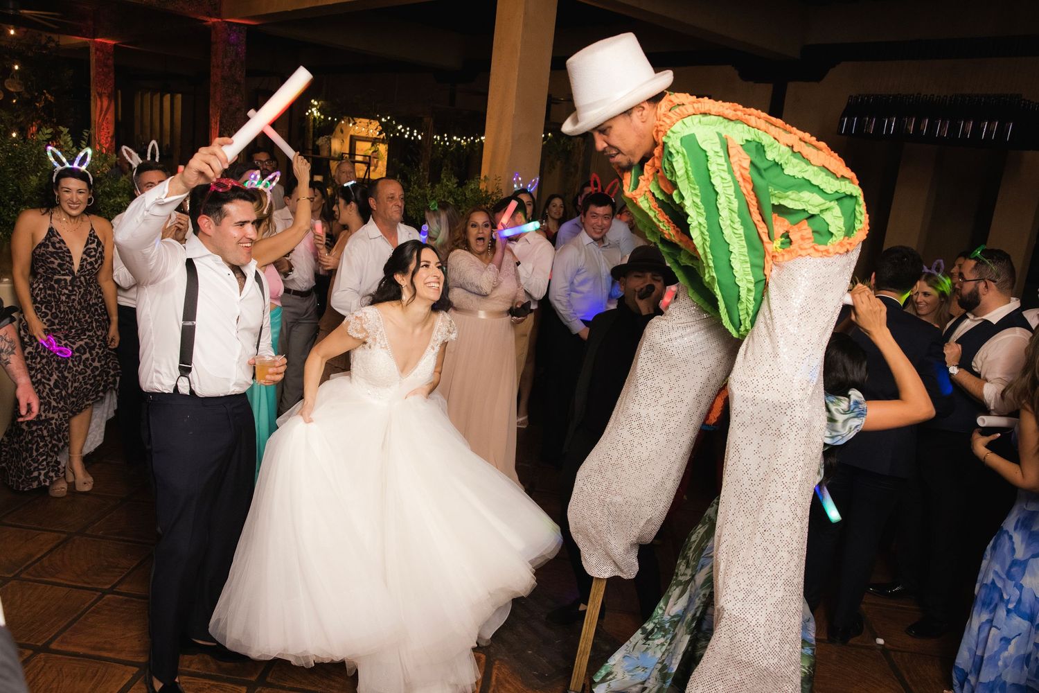 Wedding celebration on dance floor with performer on stilts entertaining guests in white and green costume during reception.