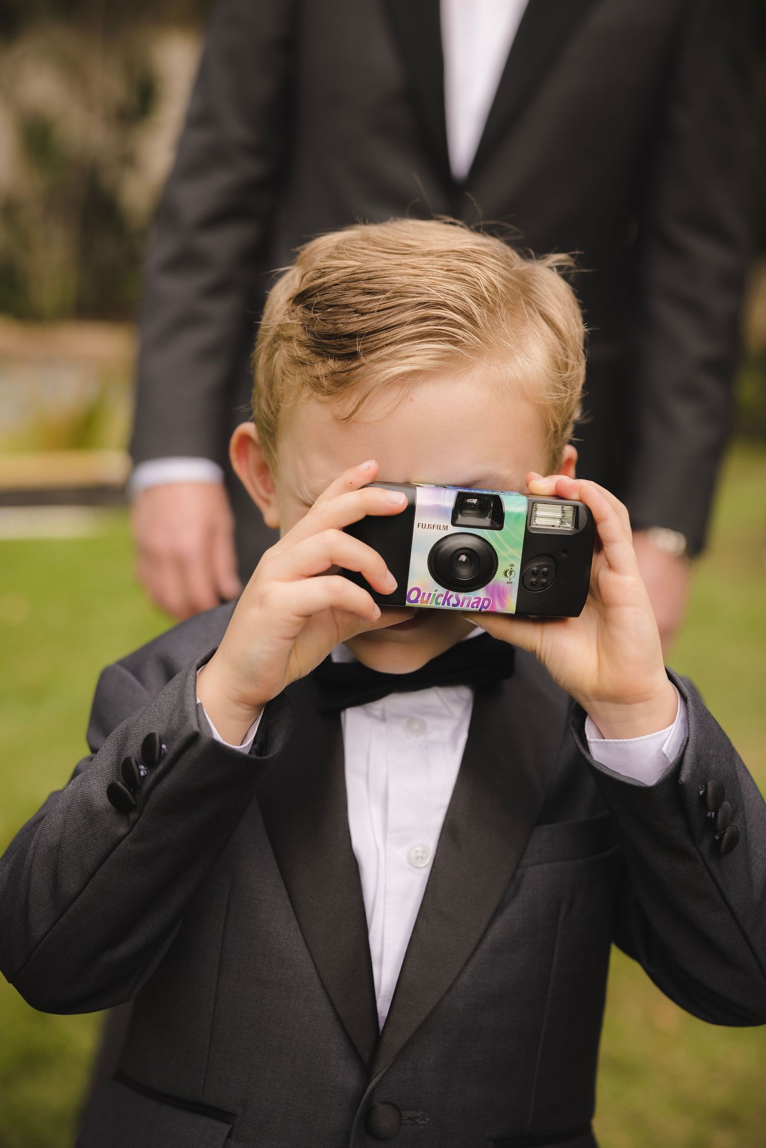 A young ring bearer in a tuxedo peeks through a disposable camera in a series of playful photos.