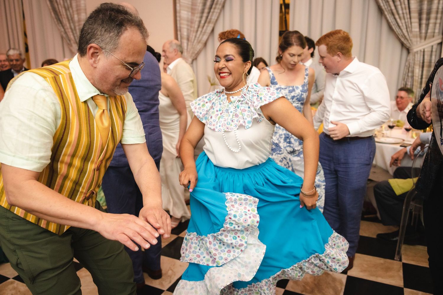 A group of people smiling and dancing together at a social gathering with beige curtains in the background.