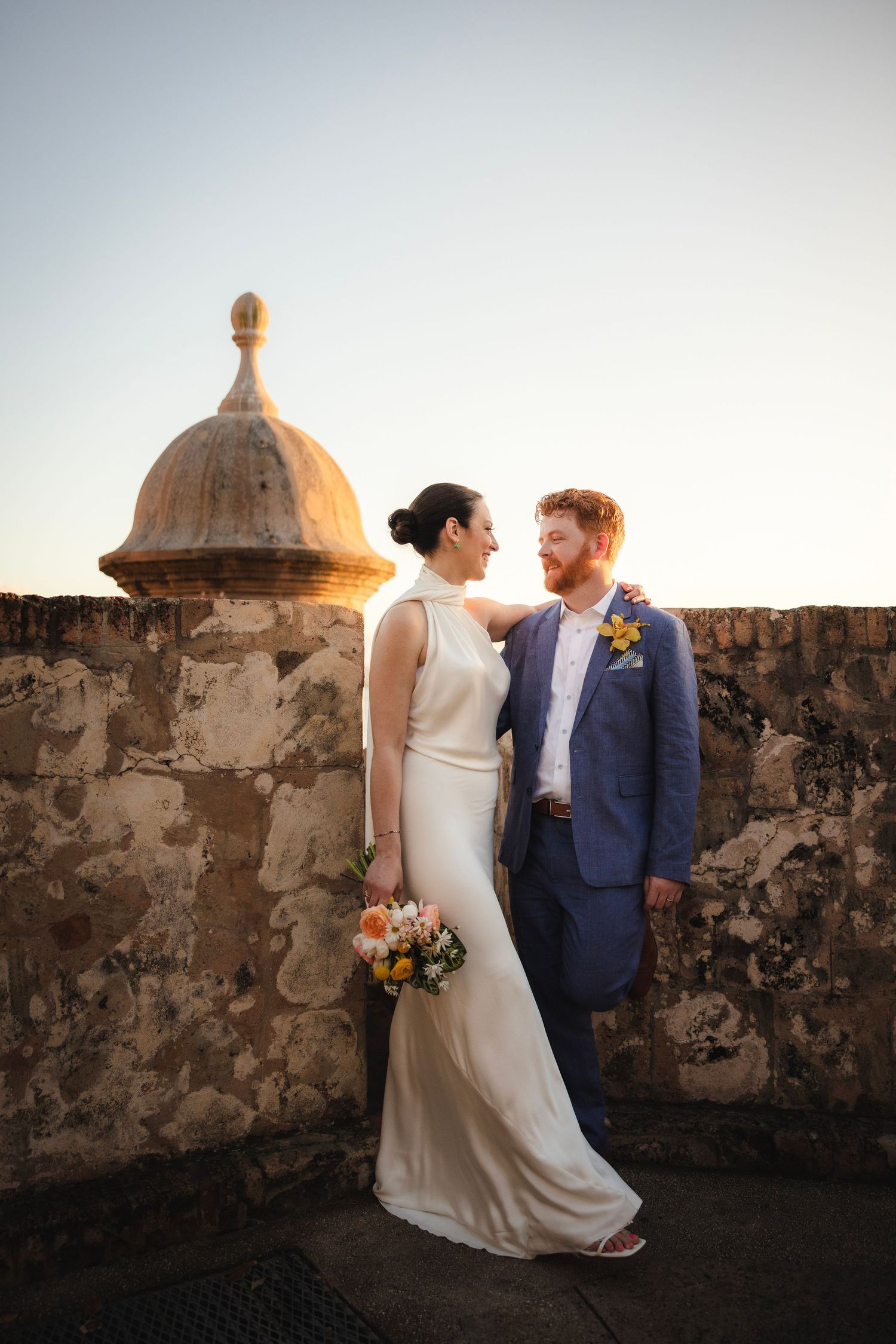 A romantic wedding photo against weathered stone walls and domed turrets at sunset.