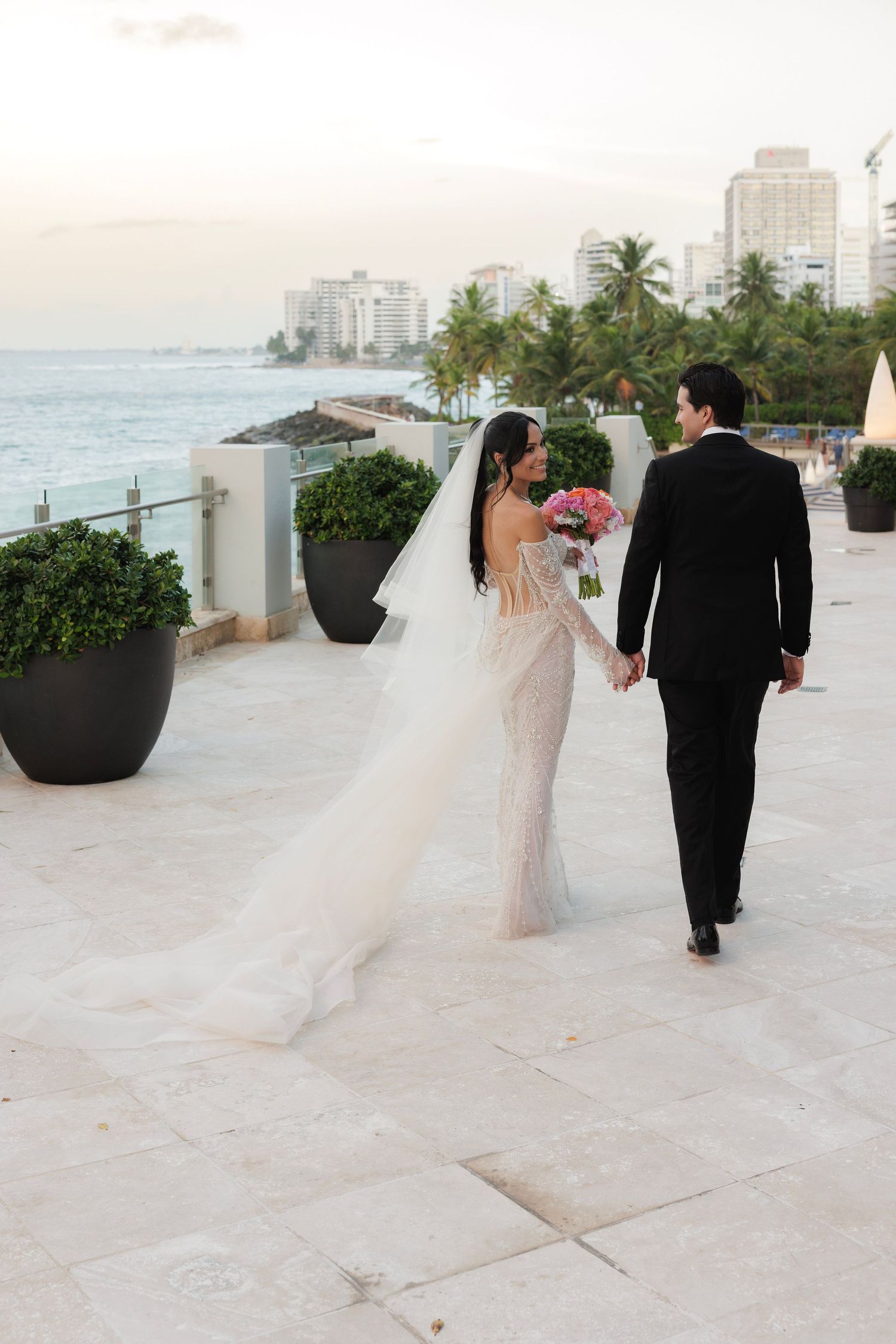 Wedding couple walks along coastal path with city skyline and palm trees in background.